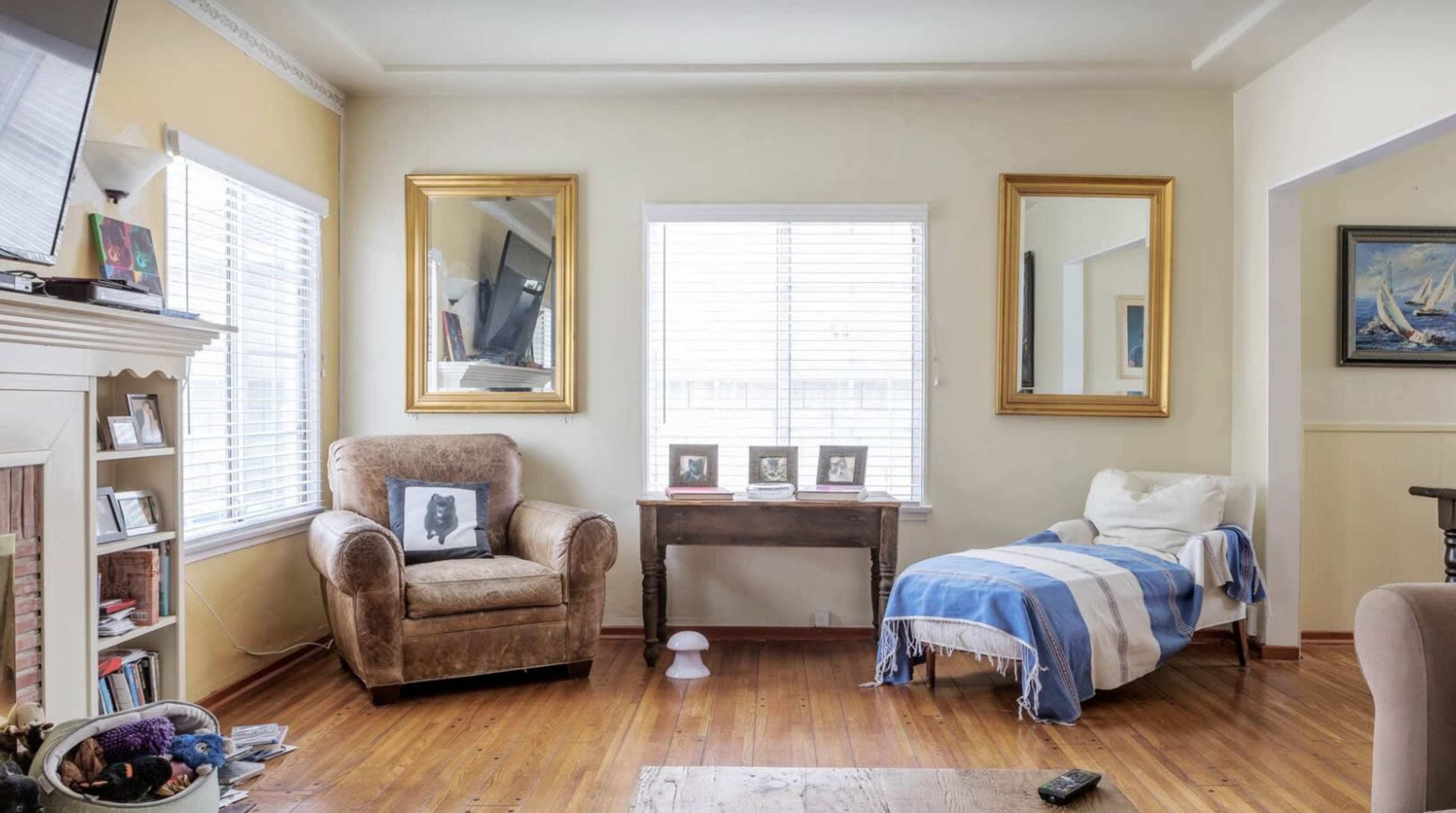 The image shows a living room with a brown chair, a white chaise lounge, two large mirrors, and framed photographs on a wooden table, all illuminated by natural light from the windows.