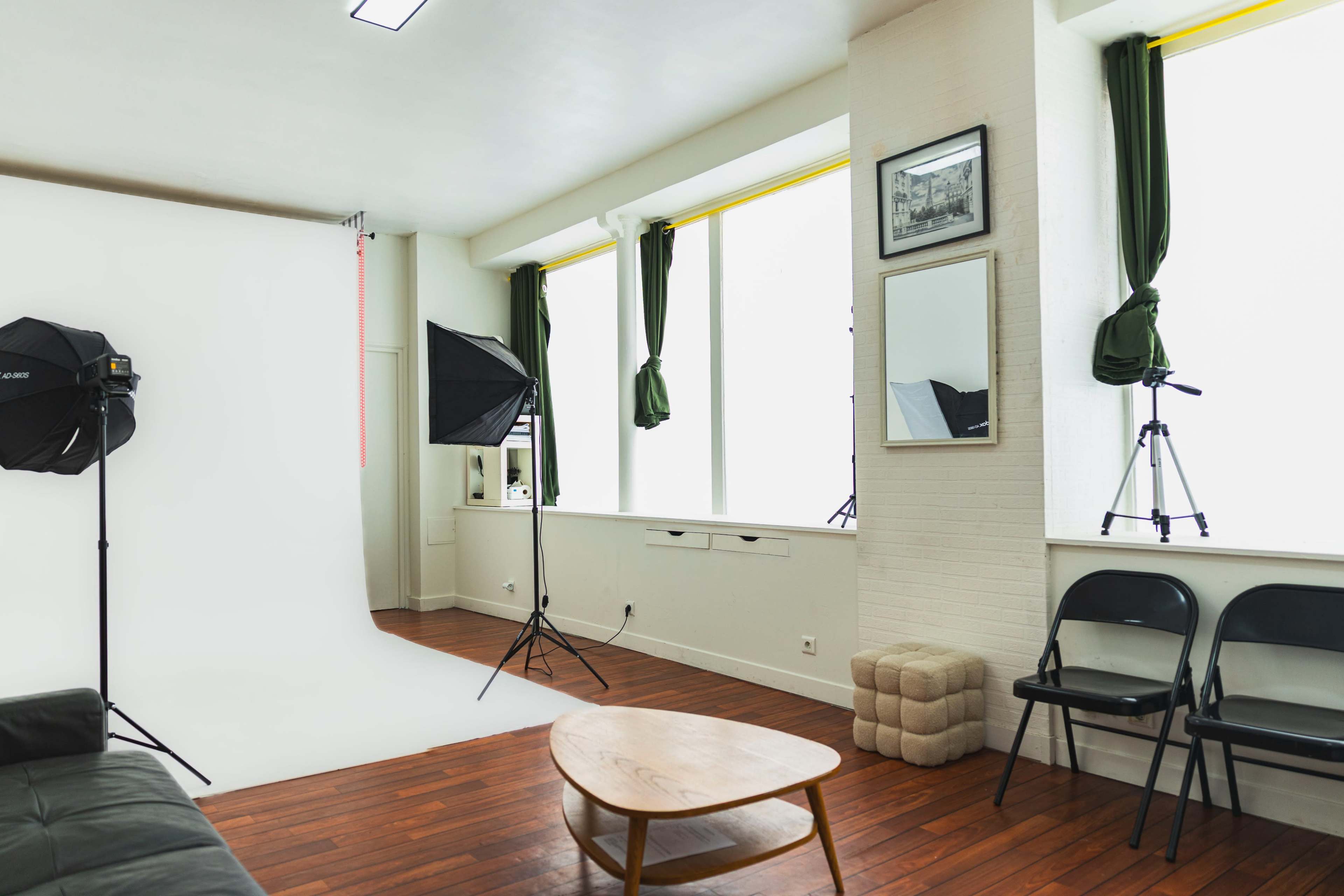 A bright photography studio features a white backdrop, two light stands, a tripod, and wooden flooring, with green curtains framing the windows.