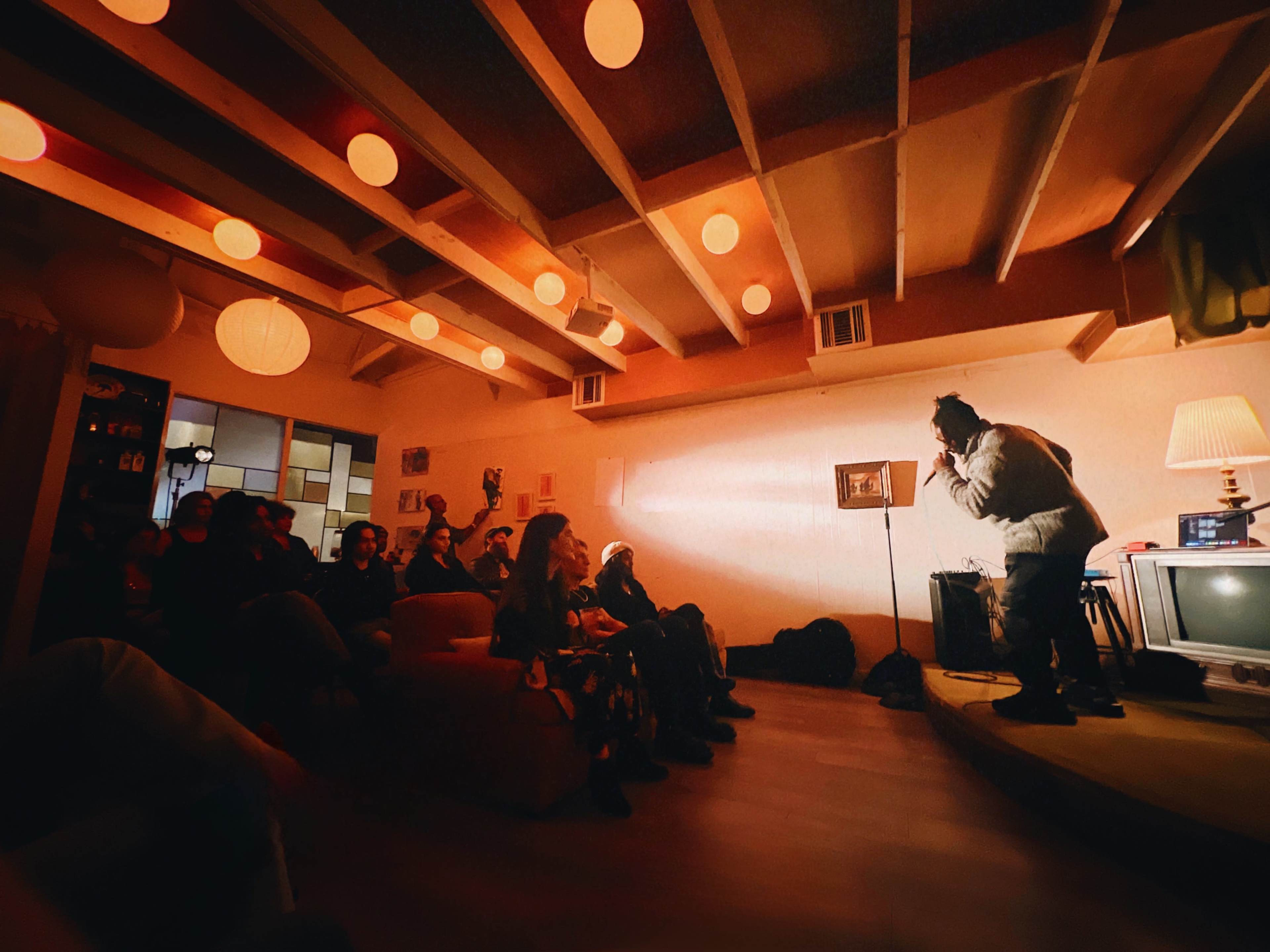 A performer stands on stage in a cozy room with a small audience seated on a couch, illuminated by soft overhead lights.