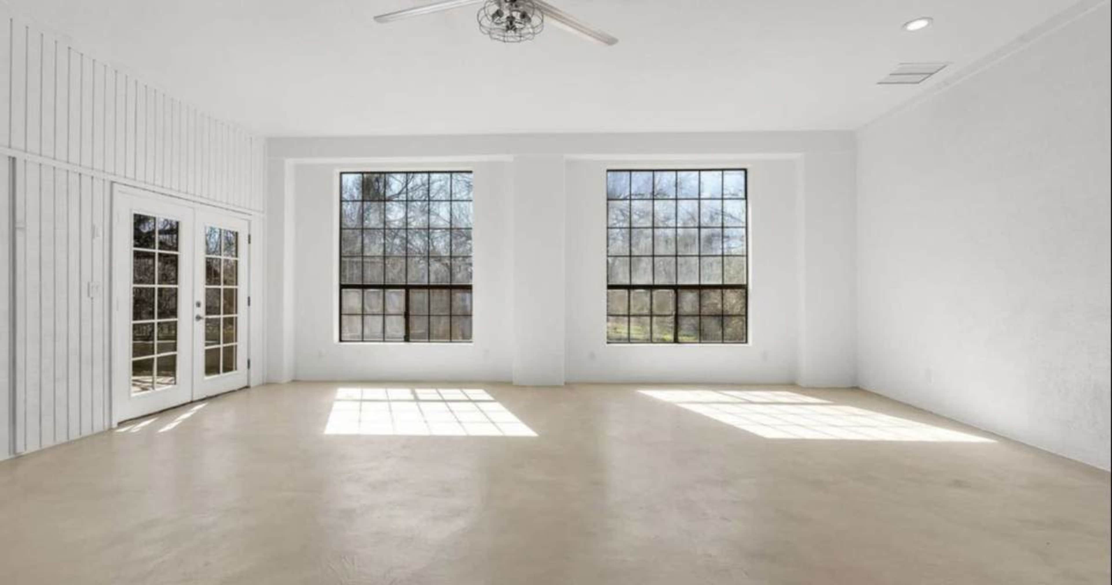 The image shows a bright, empty room with large windows on two sides and a smooth light-colored floor.