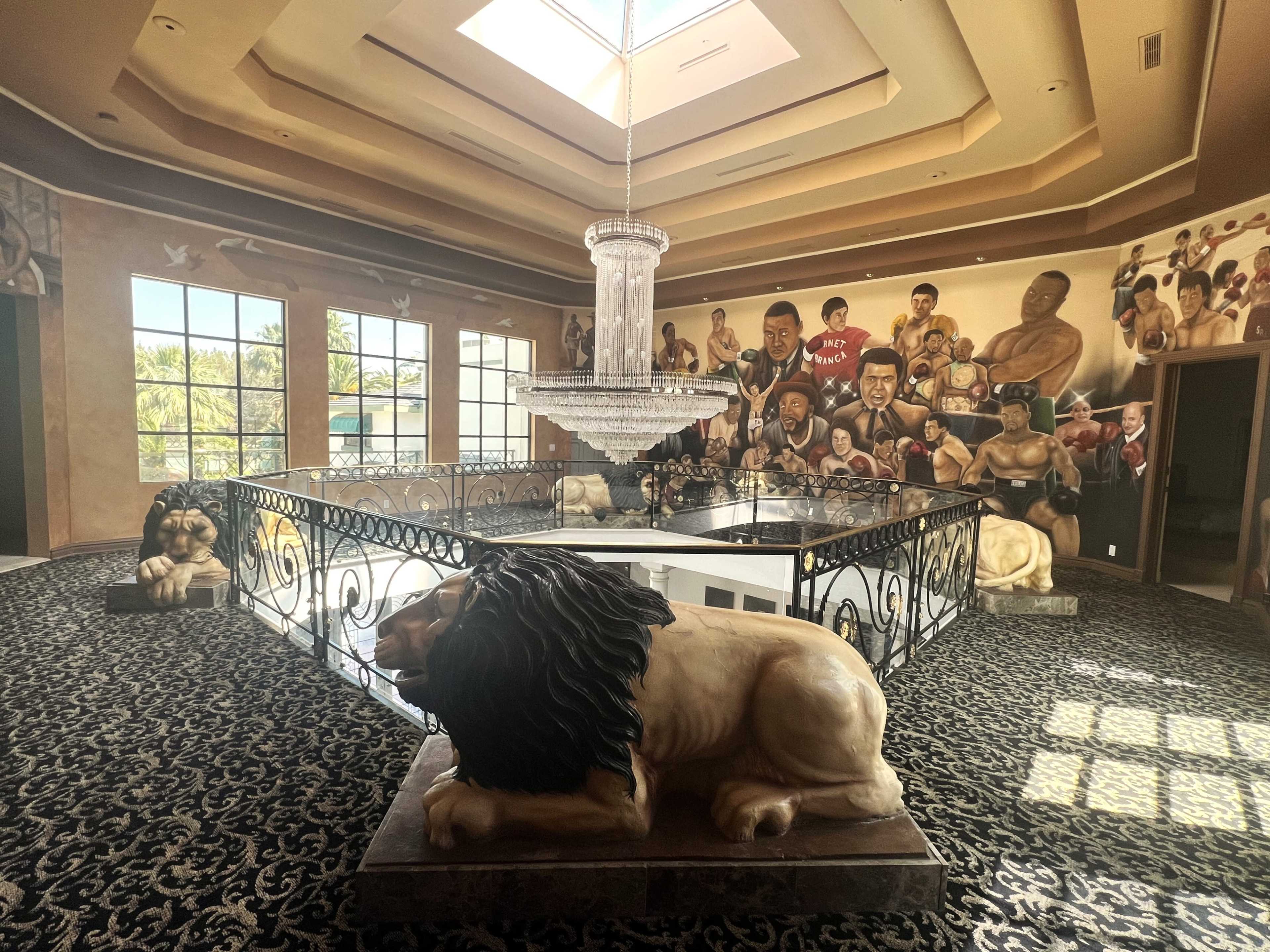 The image shows a spacious interior with a decorative railing featuring lion sculptures, a chandelier hanging from a skylight, and mural artwork on the walls.