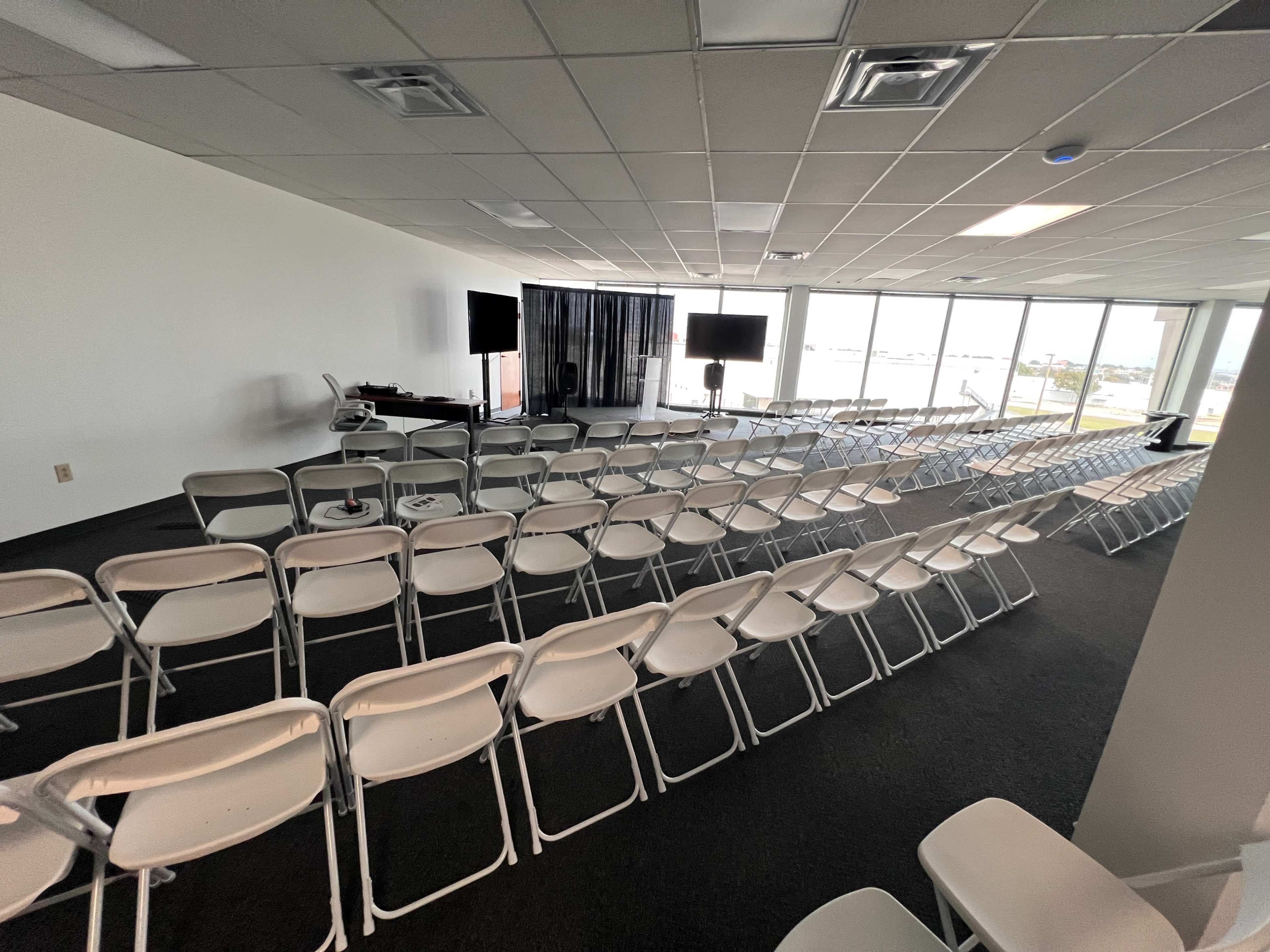 The image shows a spacious room set up with rows of white folding chairs facing a stage area, flanked by two screens.