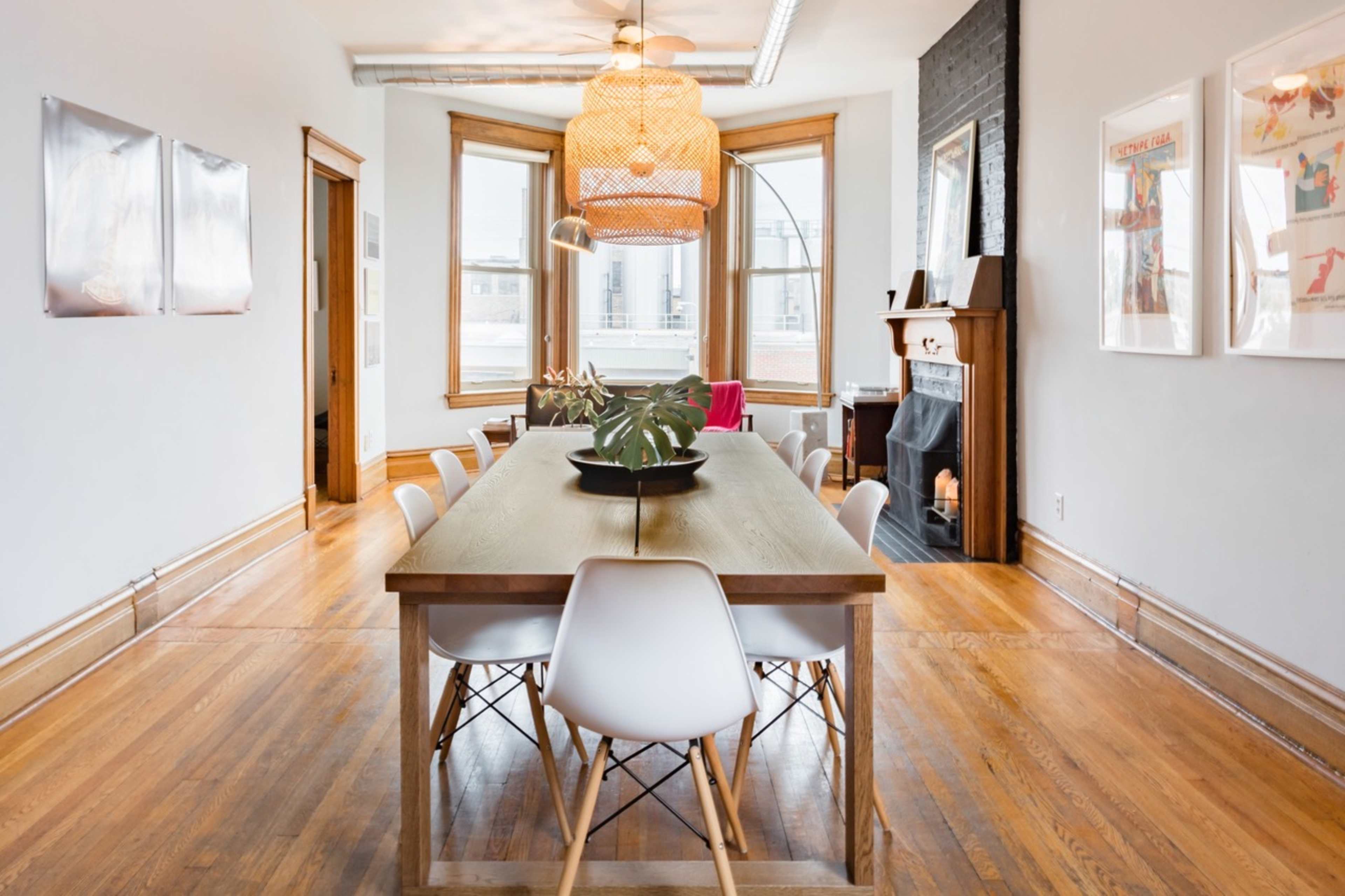 A dining area features a long wooden table surrounded by white chairs, with large windows and a decorative light fixture above.