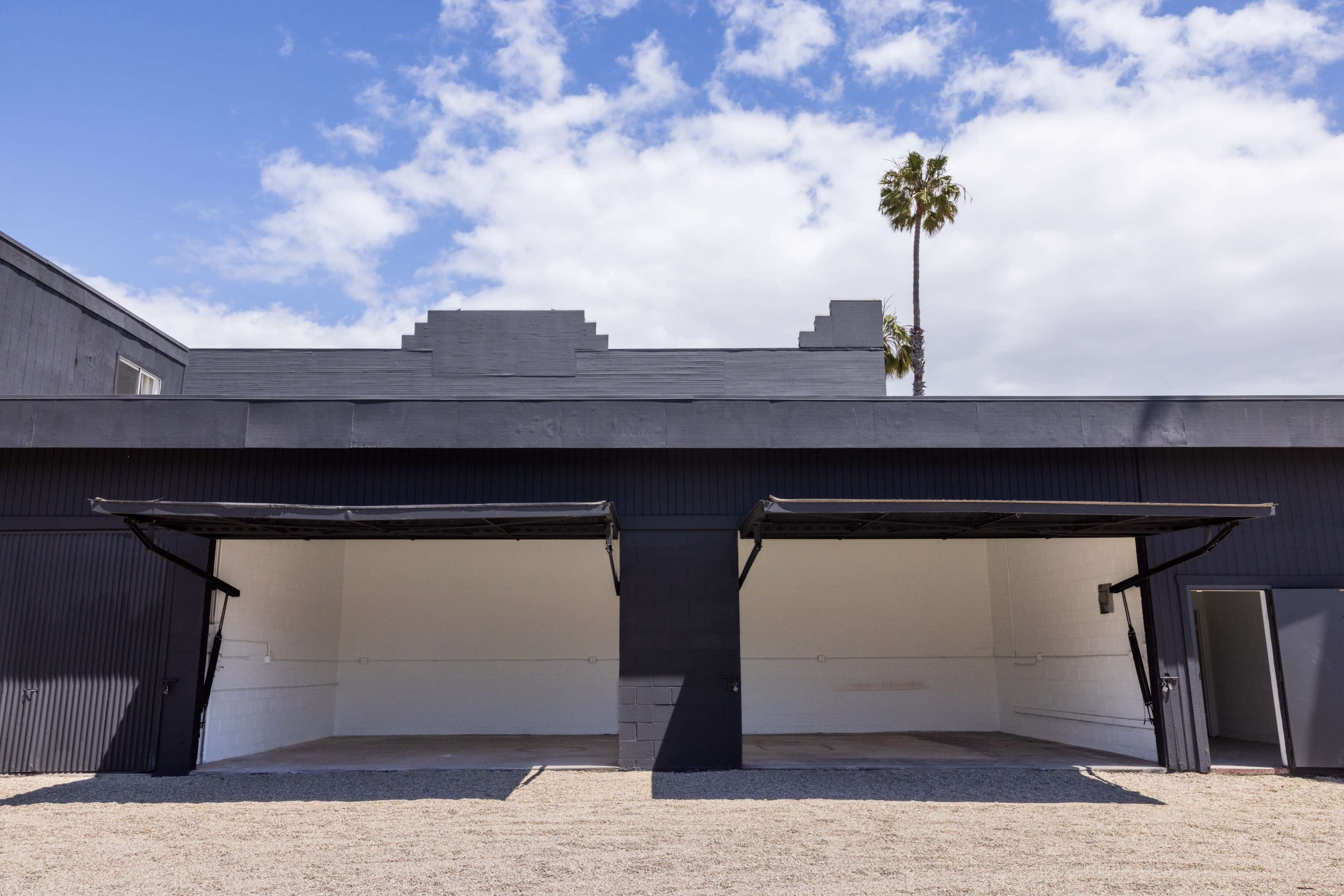 The image shows a minimalist black building with two open garage doors and a palm tree in the background under a partly cloudy sky.