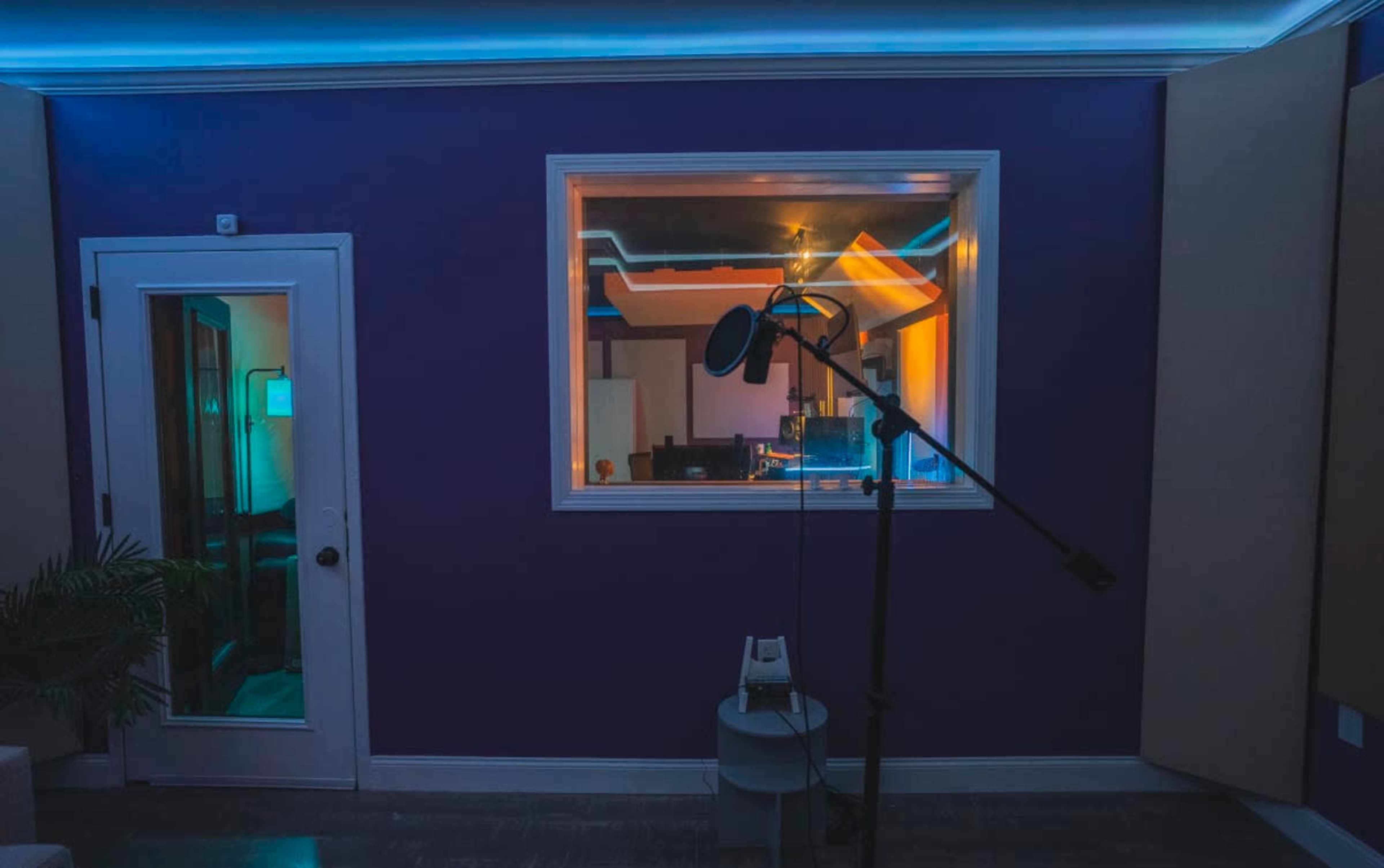 A soundproof recording studio features a microphone on a stand in front of a glass window that looks into a brightly lit control room.