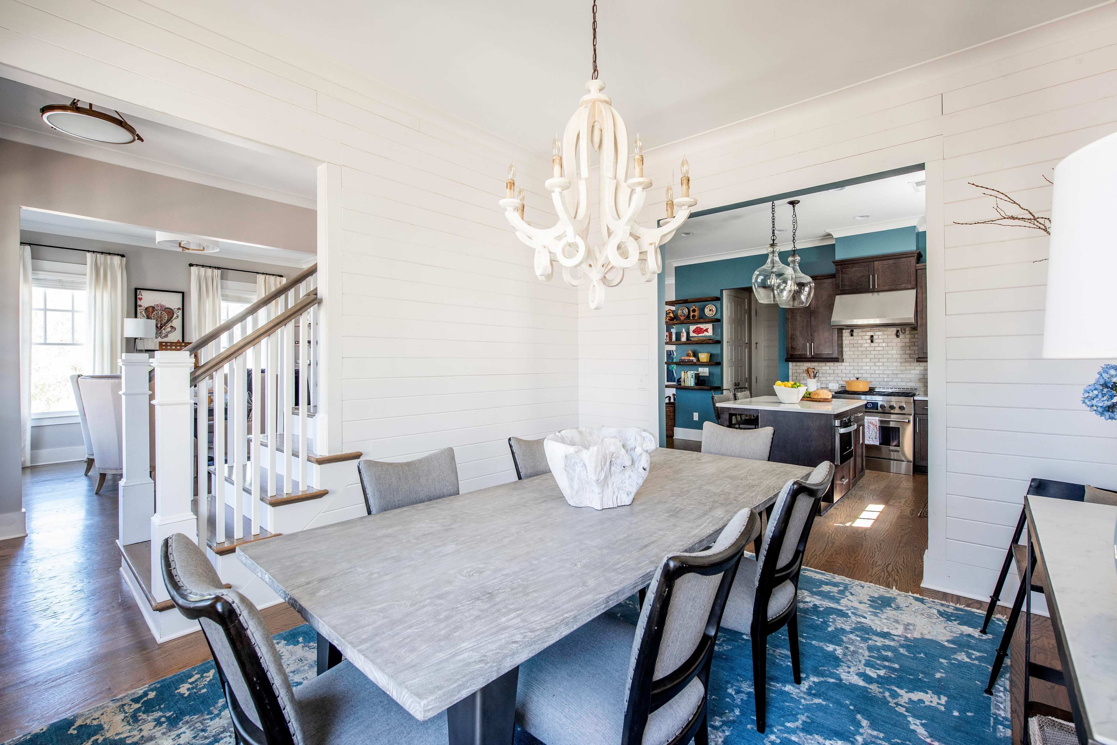 A dining room features a large table surrounded by chairs, with a chandelier overhead and a view into a kitchen area.