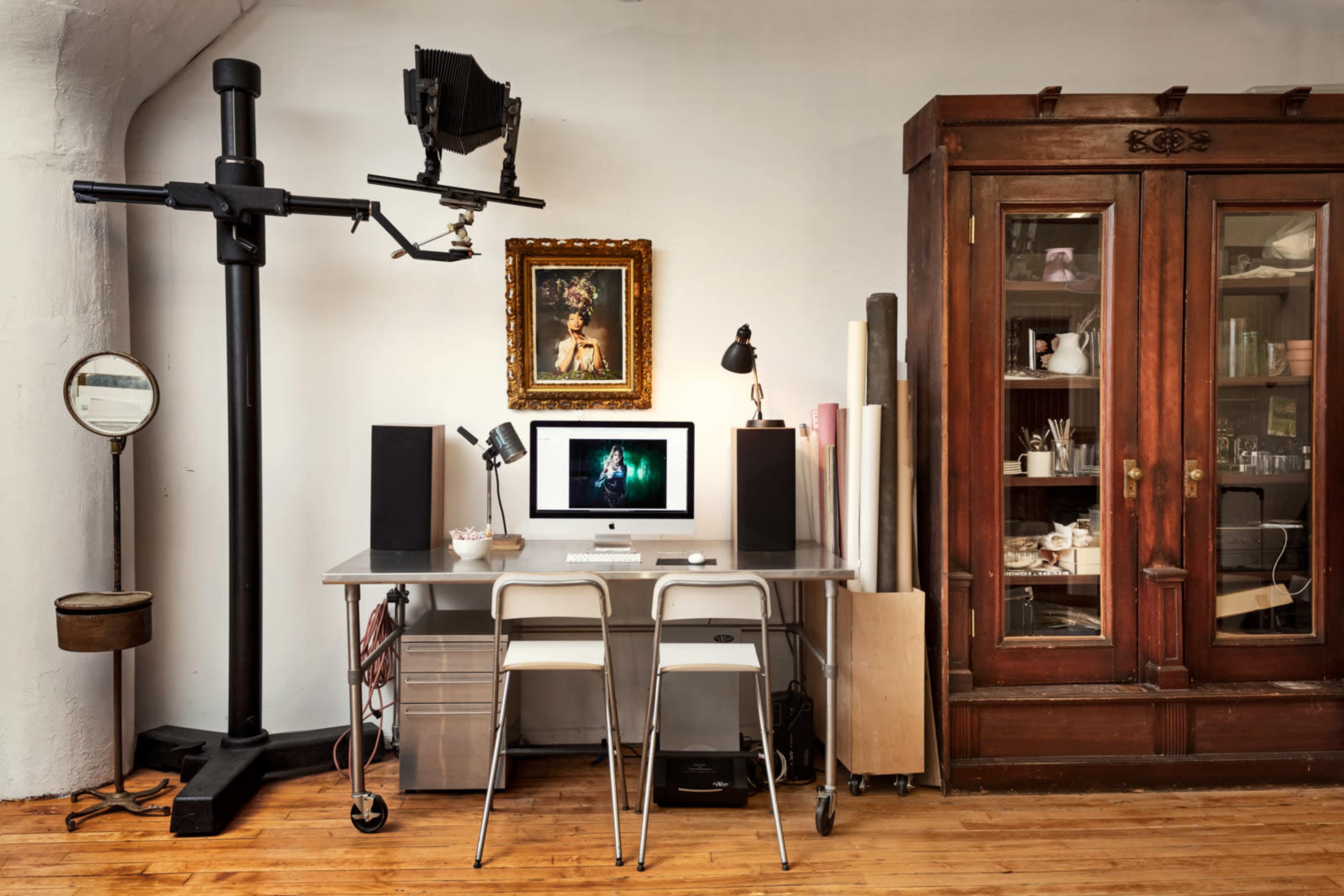 A modern workspace features a desk with an Apple computer, speakers, and two chairs, alongside a vintage wooden cabinet and decorative wall art.