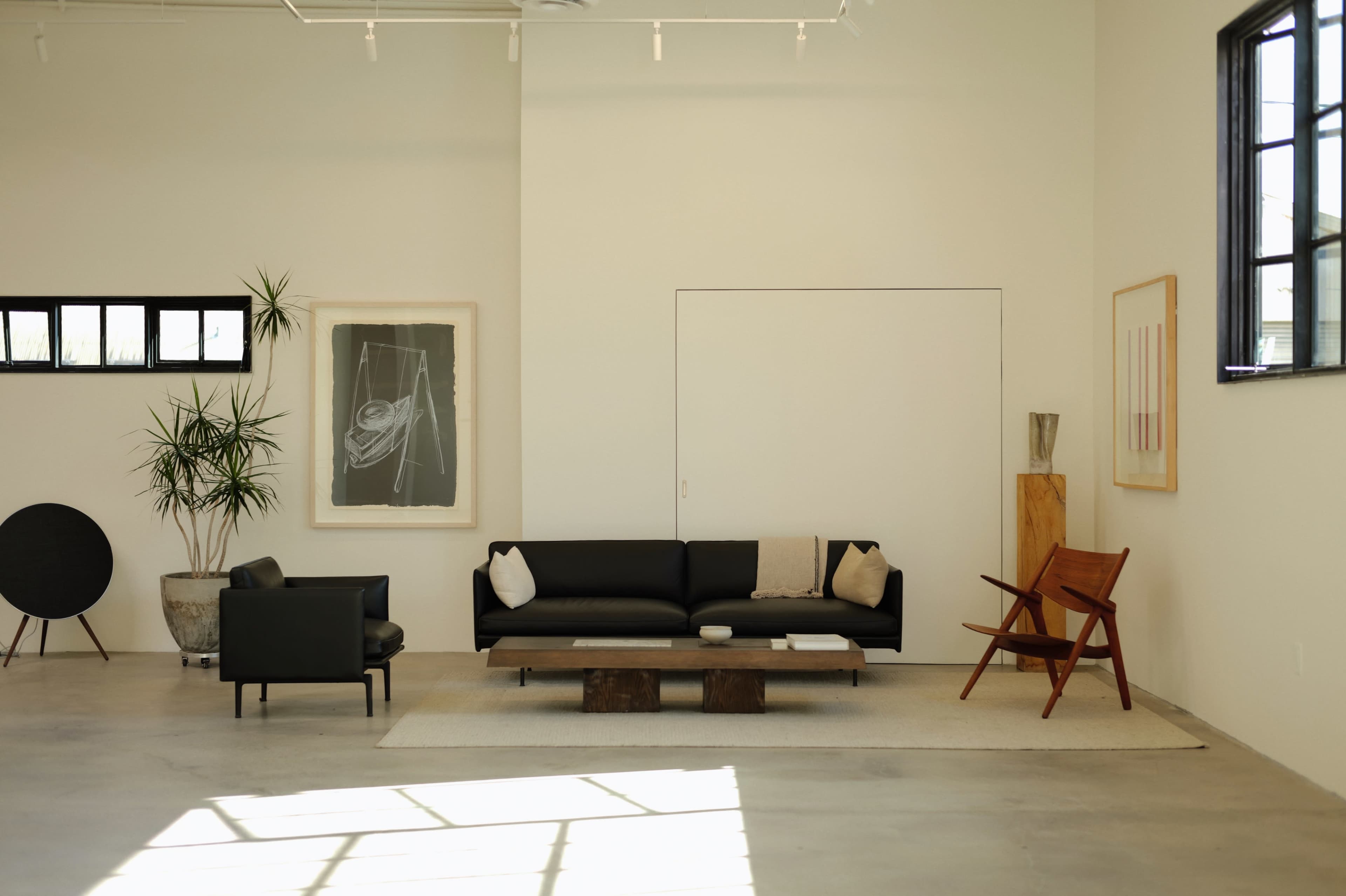 The scene depicts a minimalist living room with a black sofa, a wooden coffee table, and a single chair, complemented by a large wall art piece and potted plants.