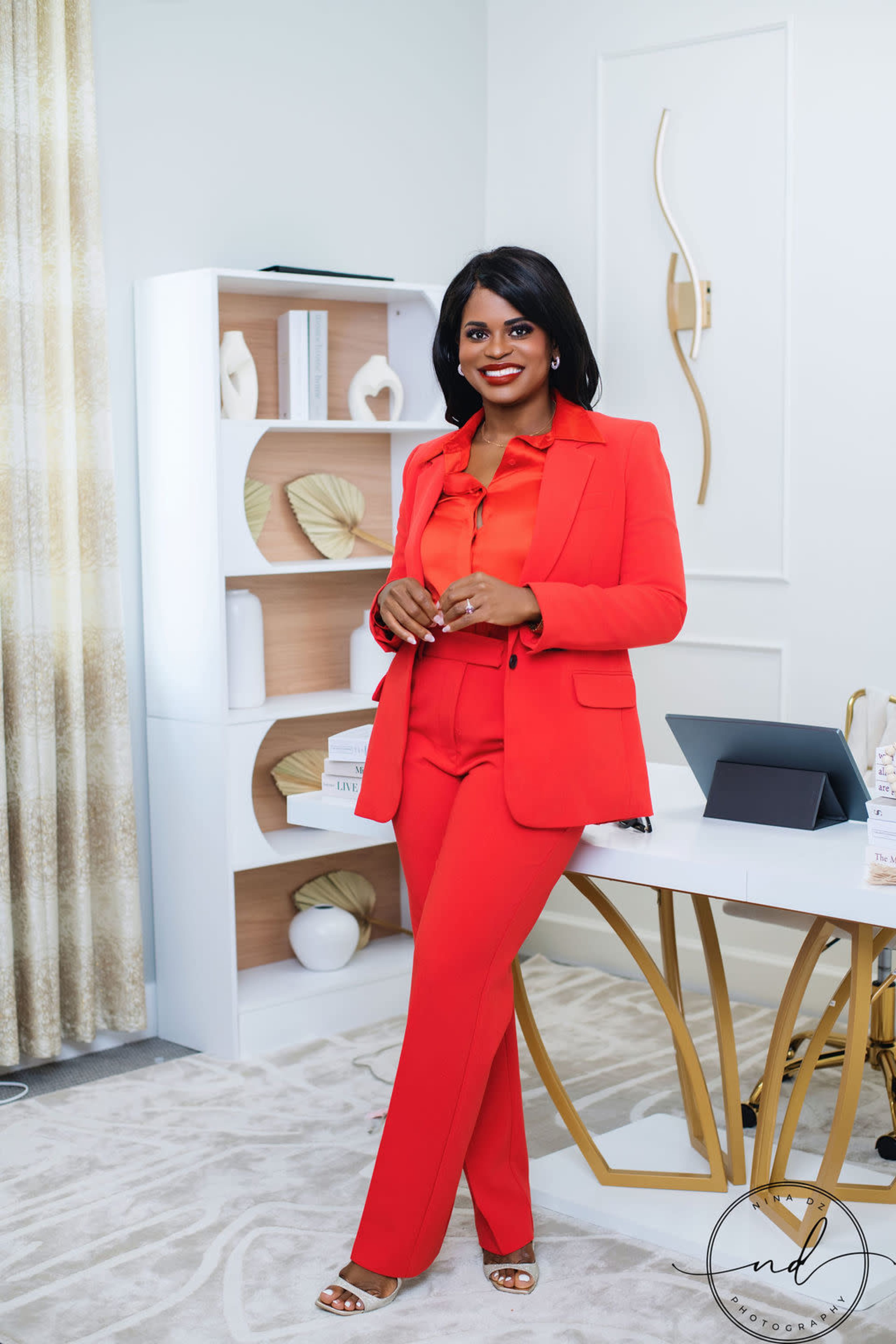 A woman in a red suit stands confidently next to a stylish desk in a modern office setting.