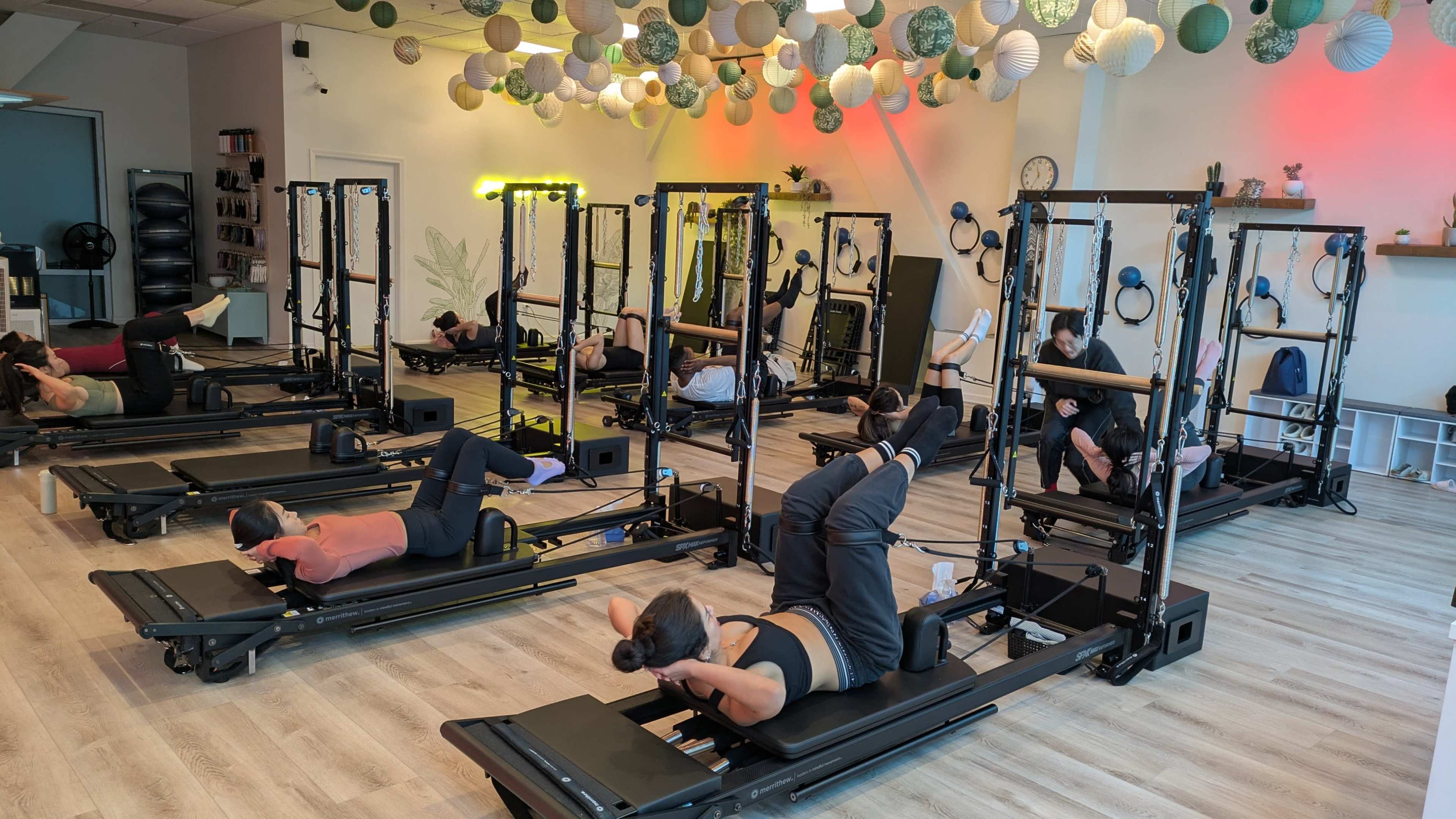 A group of people is engaged in a Pilates class using reformer machines in a brightly lit studio adorned with decorative lanterns.