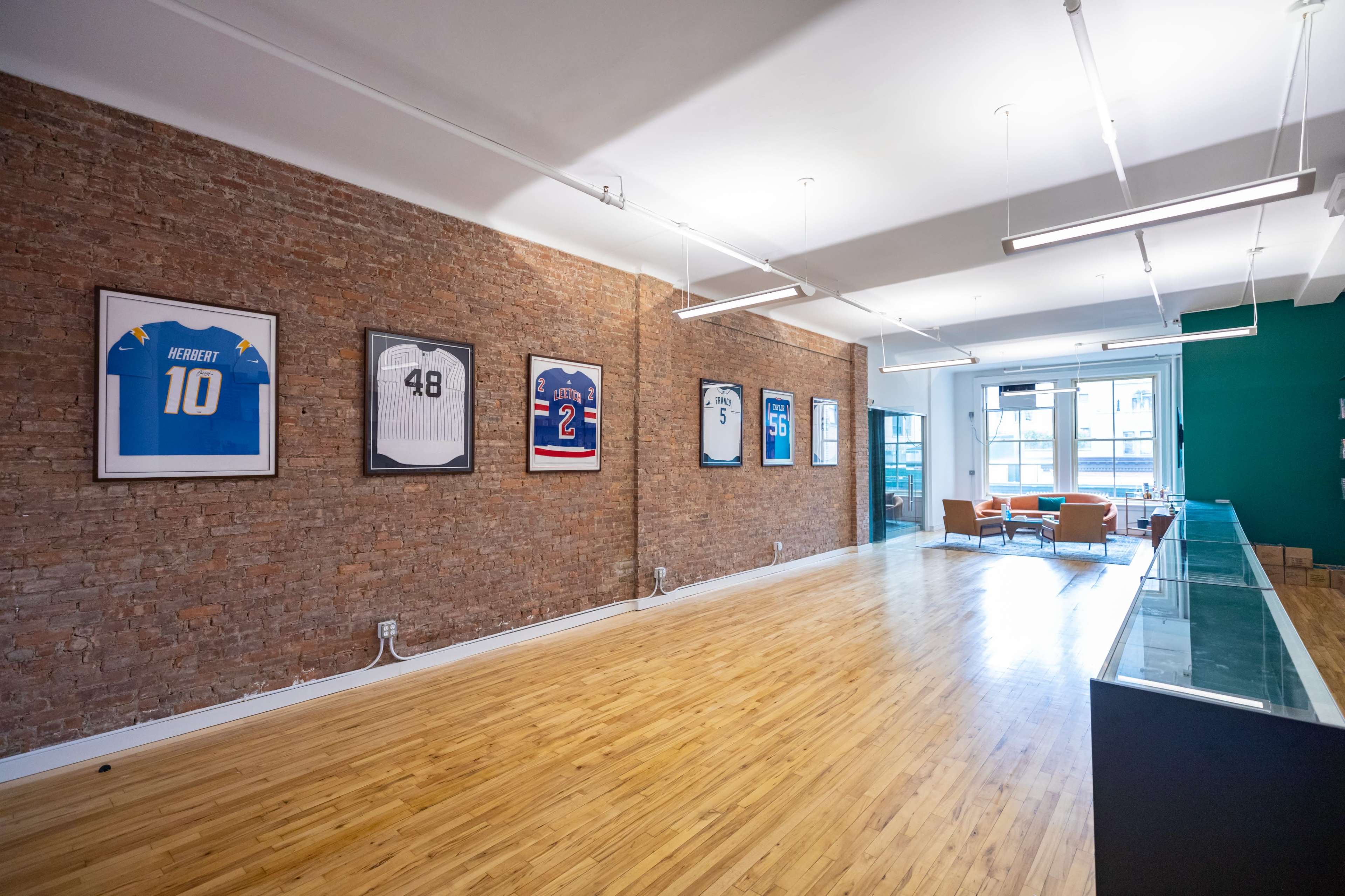 A spacious room with exposed brick walls, featuring framed sports jerseys hanging on one side and a glass display case along the other, with wooden flooring and a seating area in the corner.