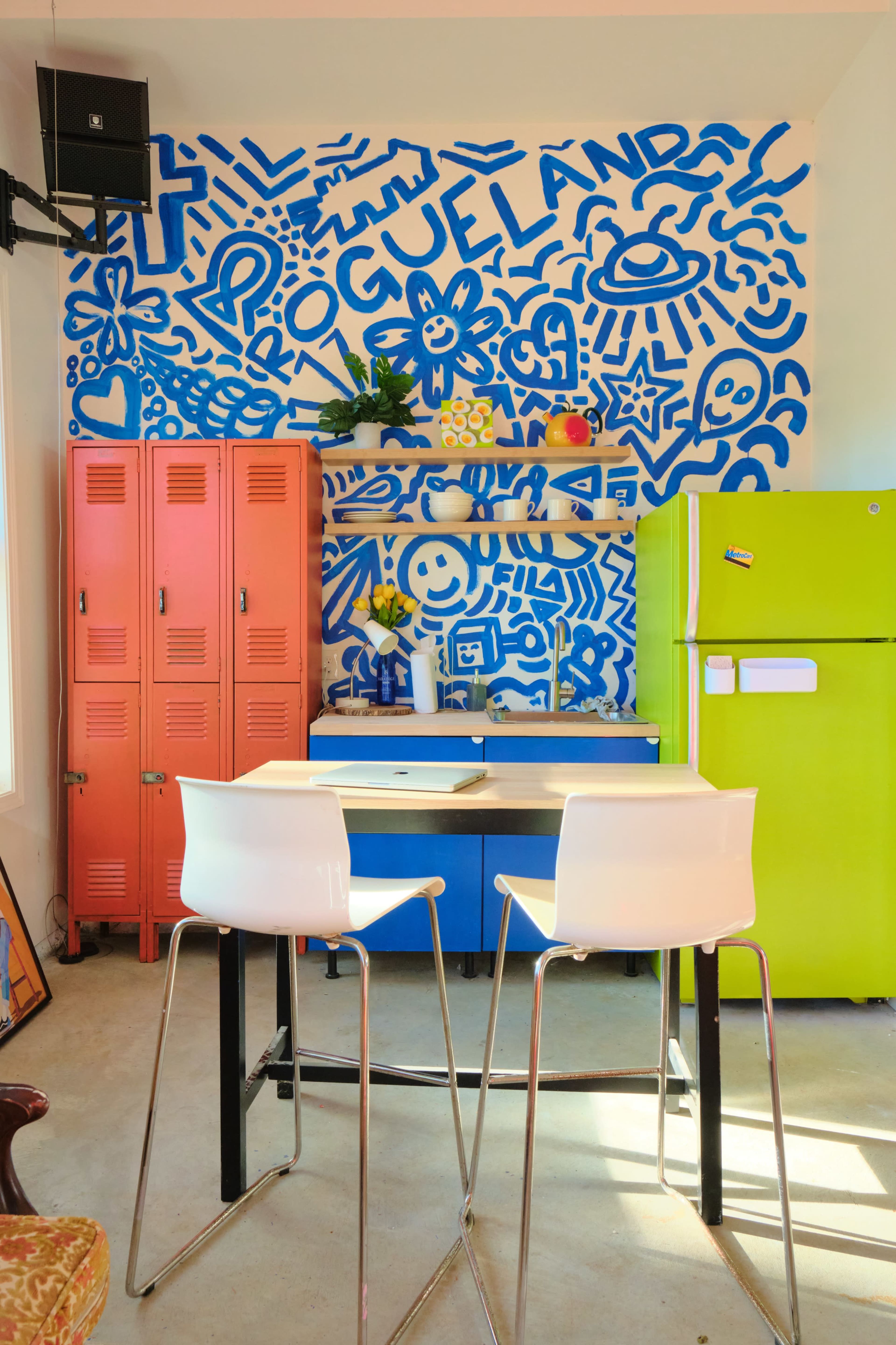 The image shows a colorful kitchen with blue graffiti-style wall art, bright red lockers, a green refrigerator, and a table with two white bar stools.