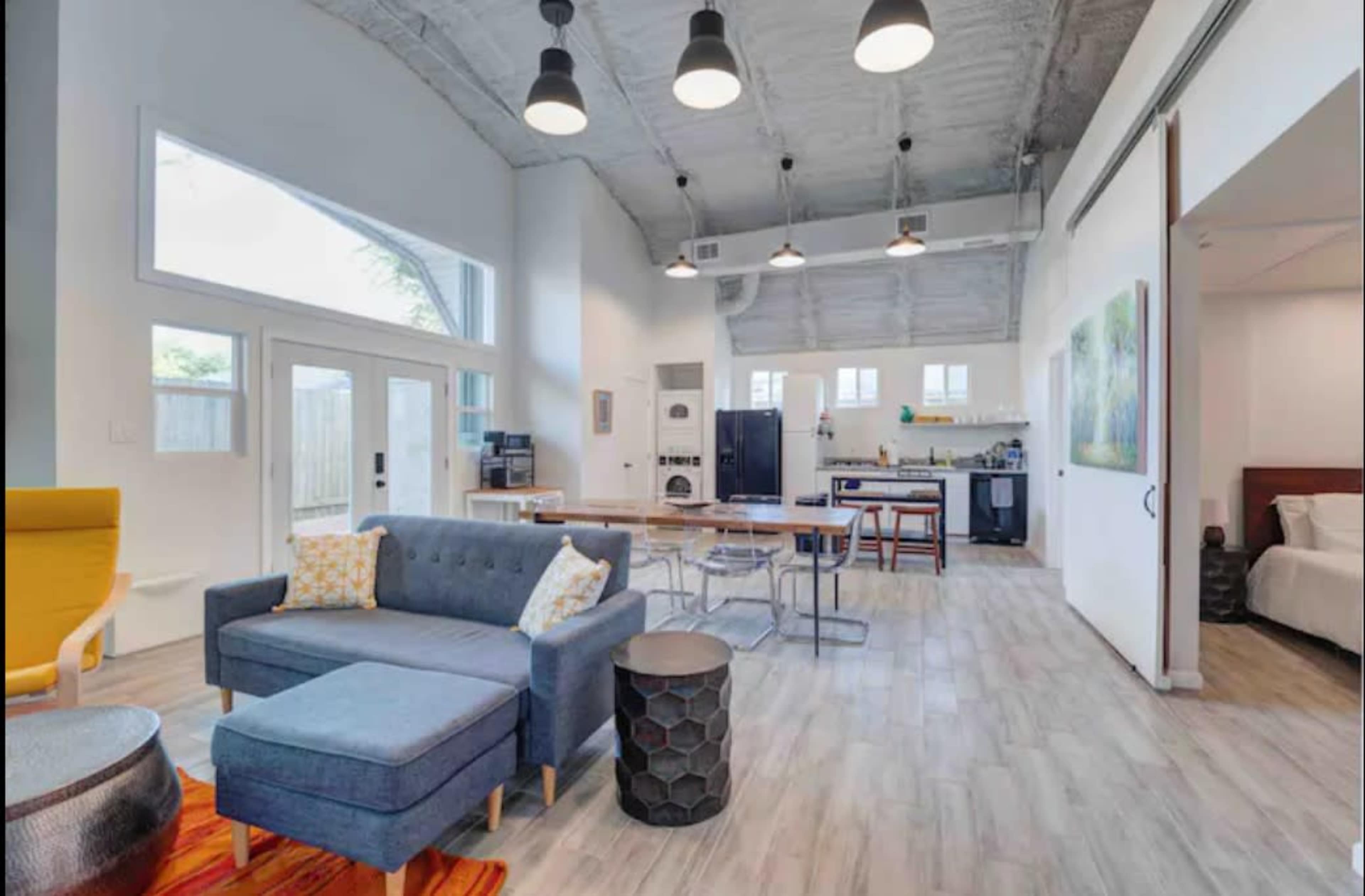A spacious, modern living area with a gray couch, a yellow accent chair, dining tables, and a kitchen in an open floor plan.