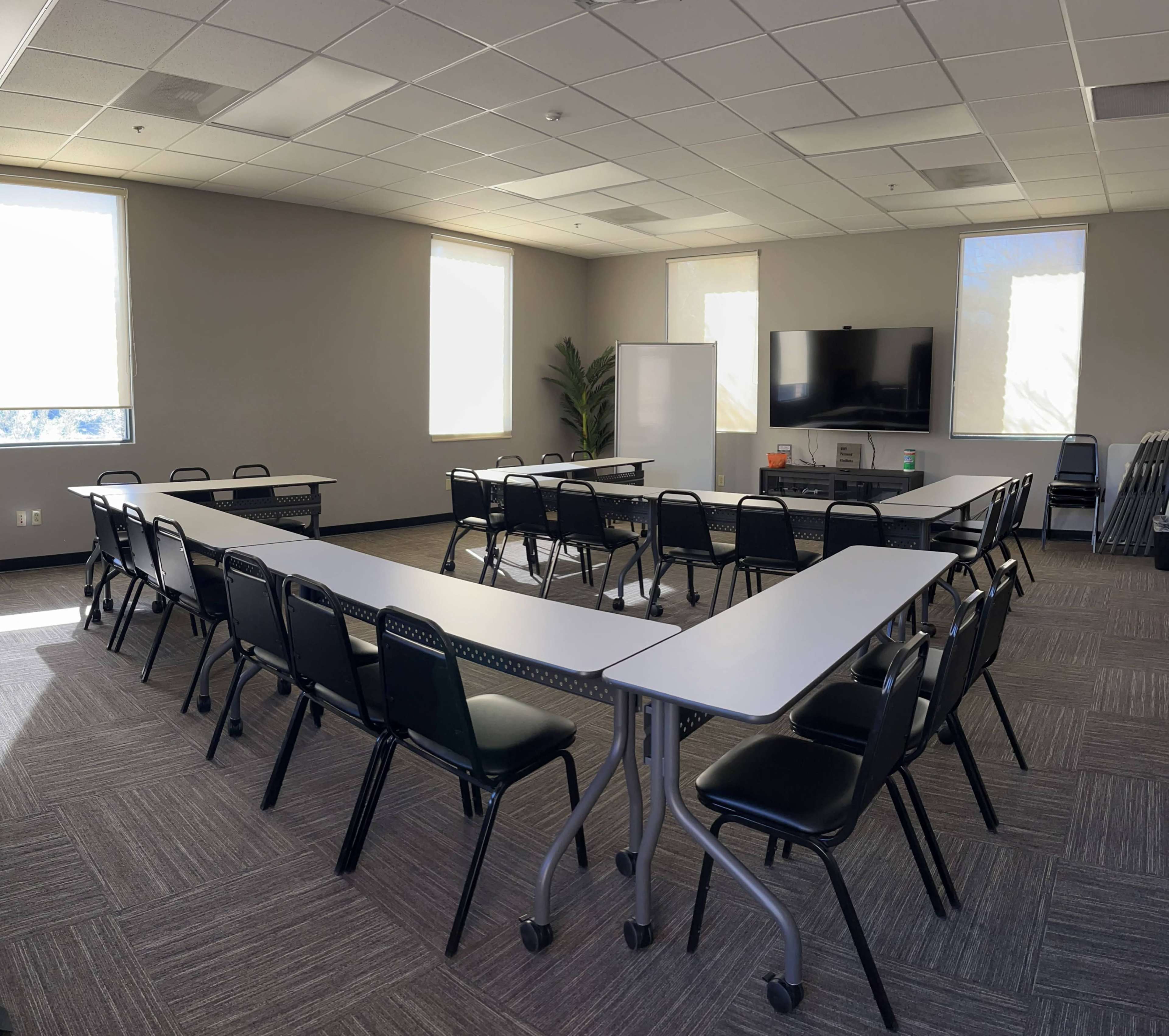 The image shows a modern classroom arranged with a U-shaped seating layout, featuring multiple tables and chairs, a whiteboard, and a TV monitor in the front.