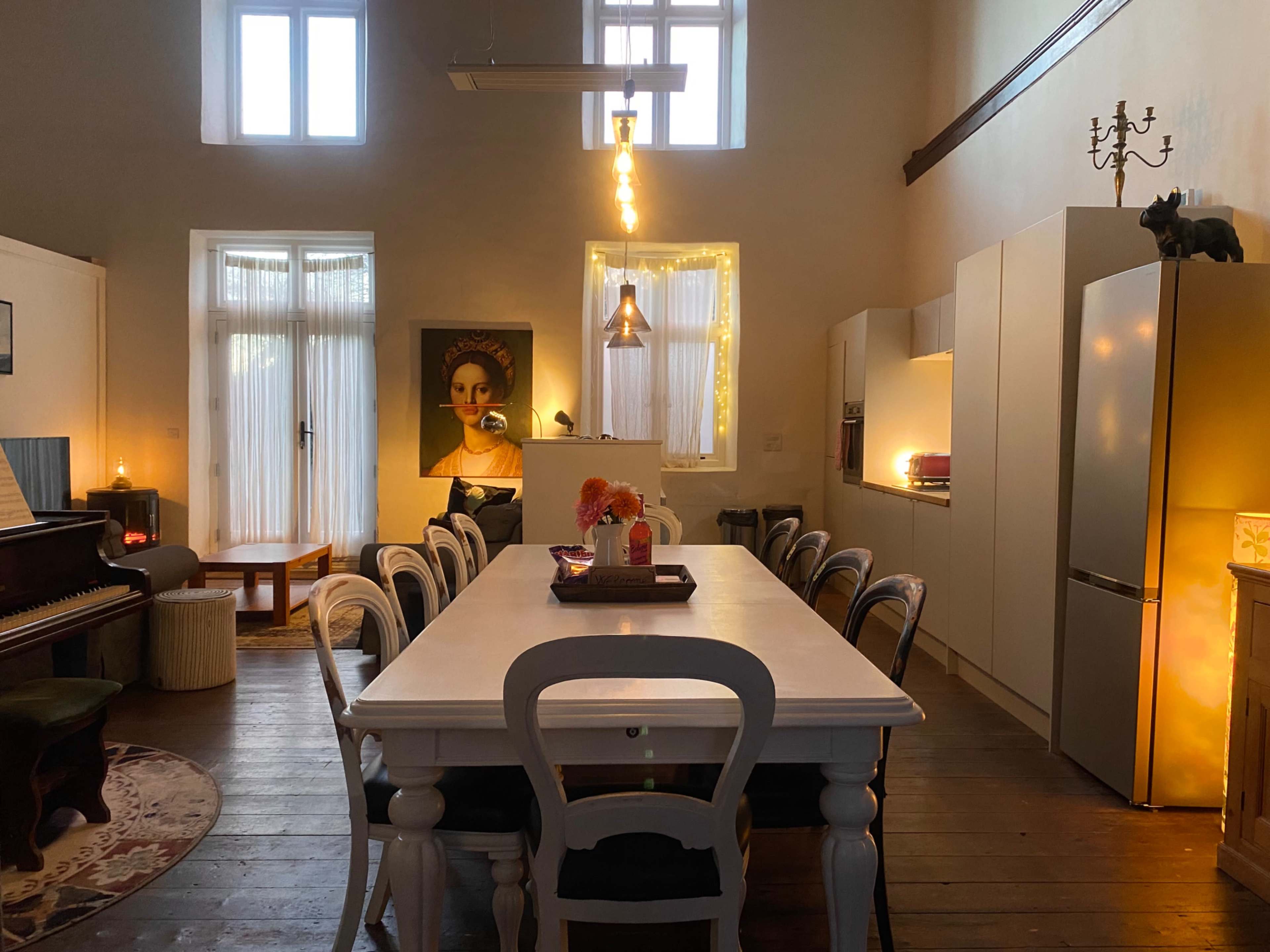 The image shows a spacious, well-lit dining area featuring a long white table, modern kitchen appliances, a piano, and decorative artwork on the walls.