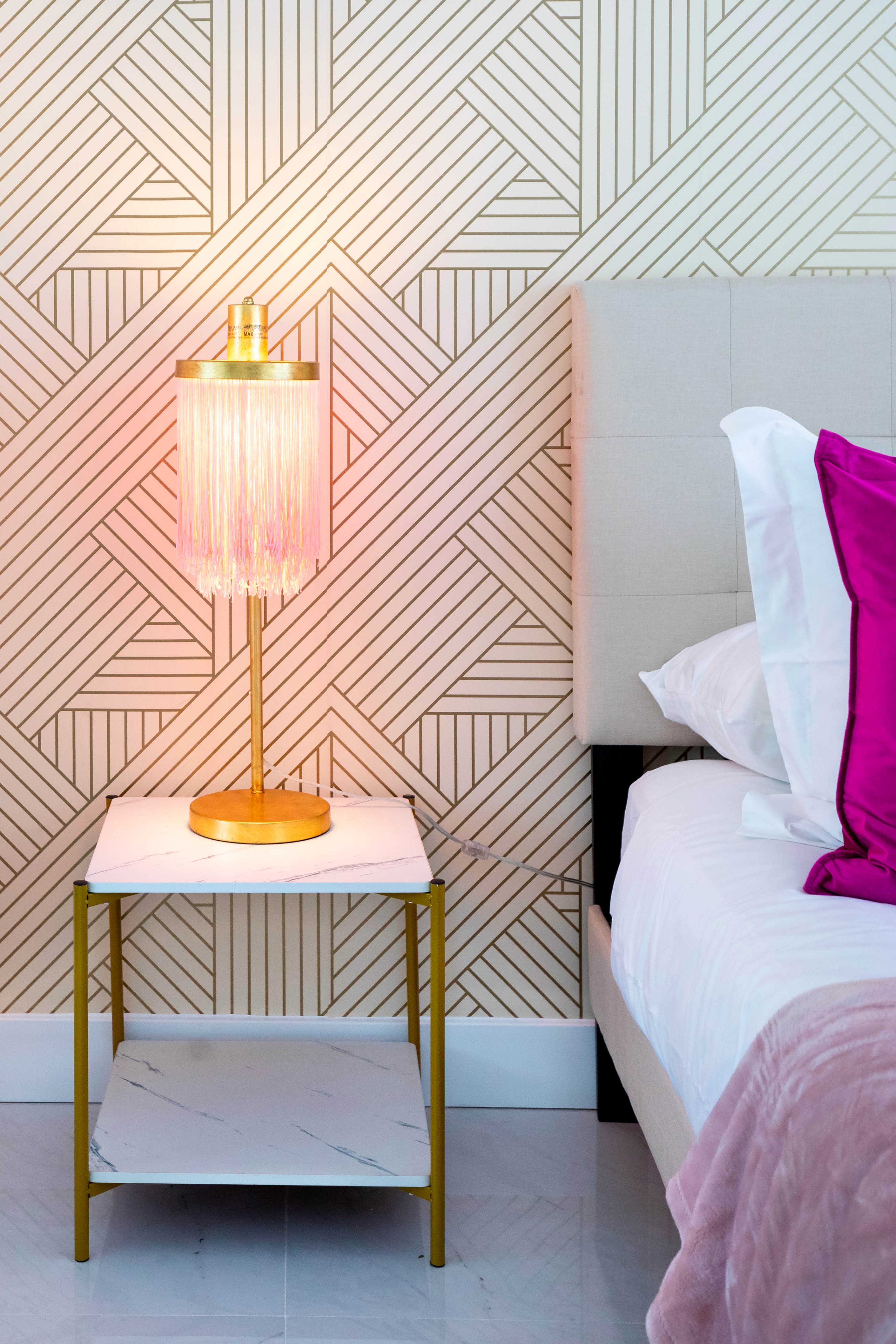 A bedside table with a fringed lamp stands next to a neatly made bed against a patterned wallpaper backdrop.