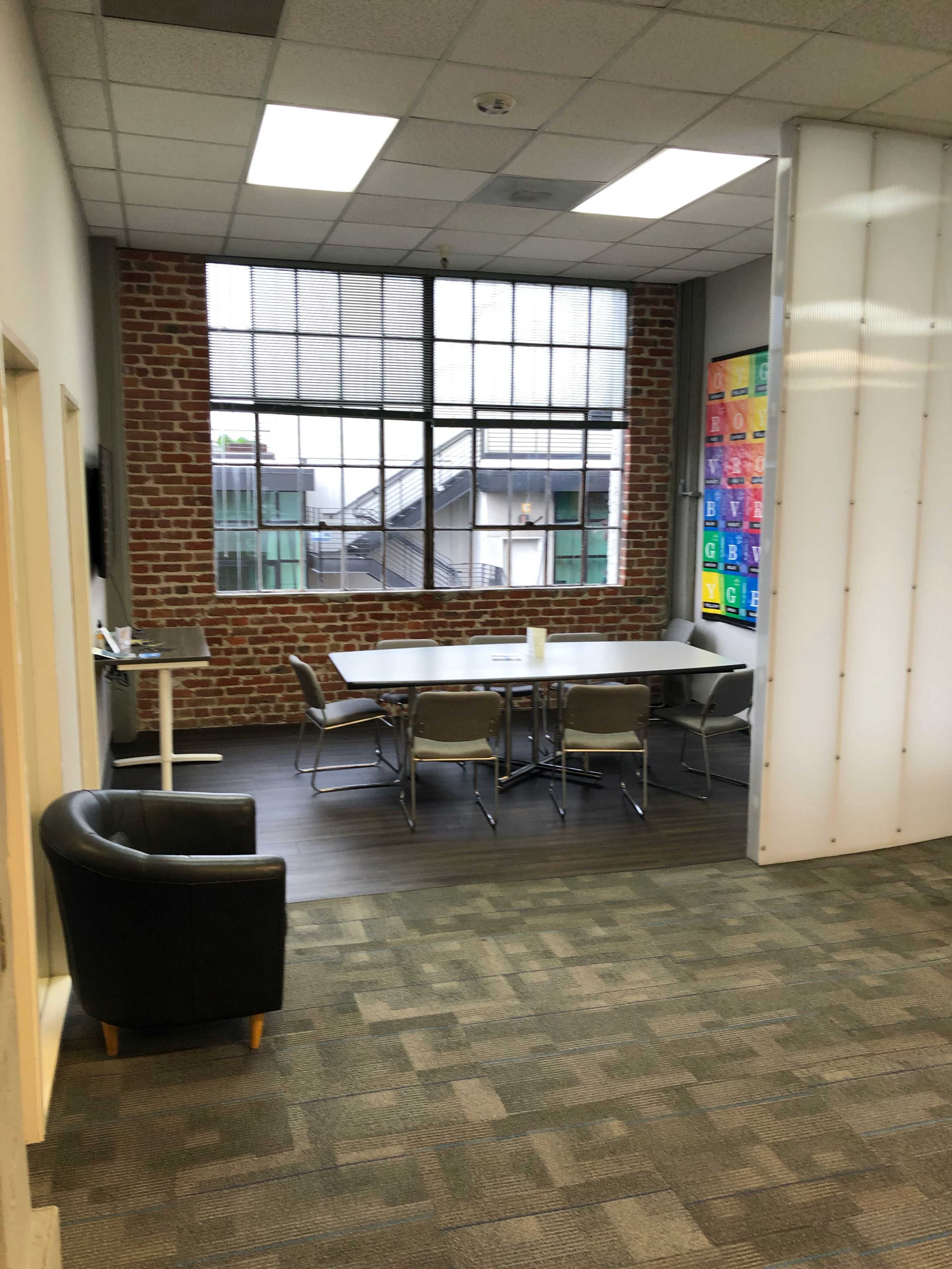 A meeting room with a rectangular table surrounded by chairs, a brick wall, large windows, and a separate seating area featuring a black chair.