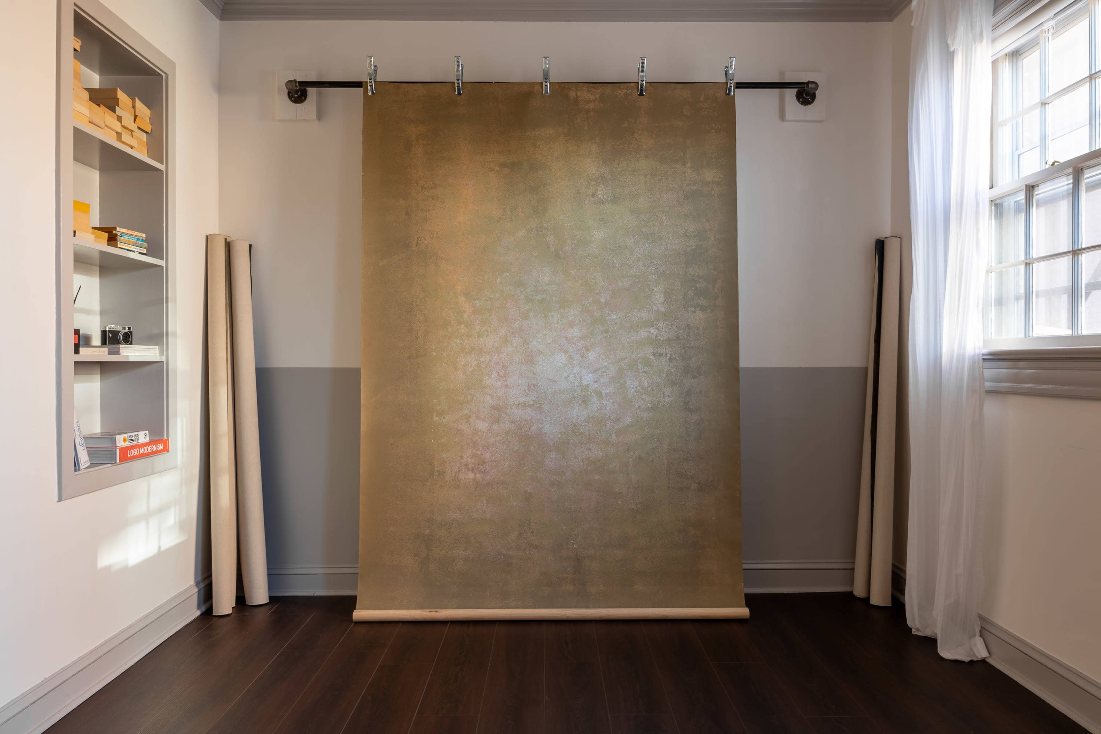 The image shows a minimalist room with a large rollable backdrop hung on a track, flanked by rolled materials and a shelf containing various items.