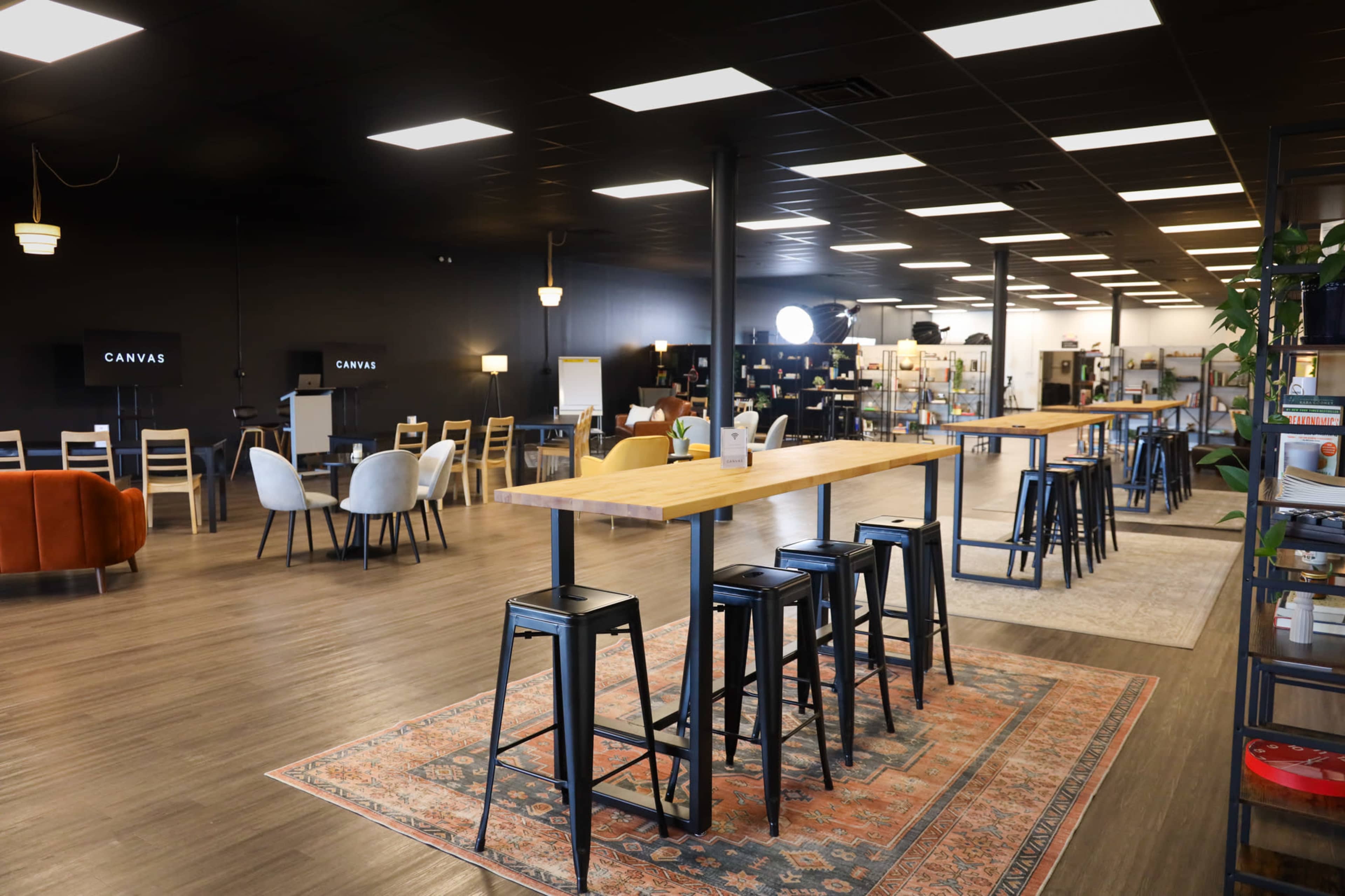 The image shows a modern co-working space with a long wooden table surrounded by black stools, and various seating areas with chairs and tables in a well-lit environment.