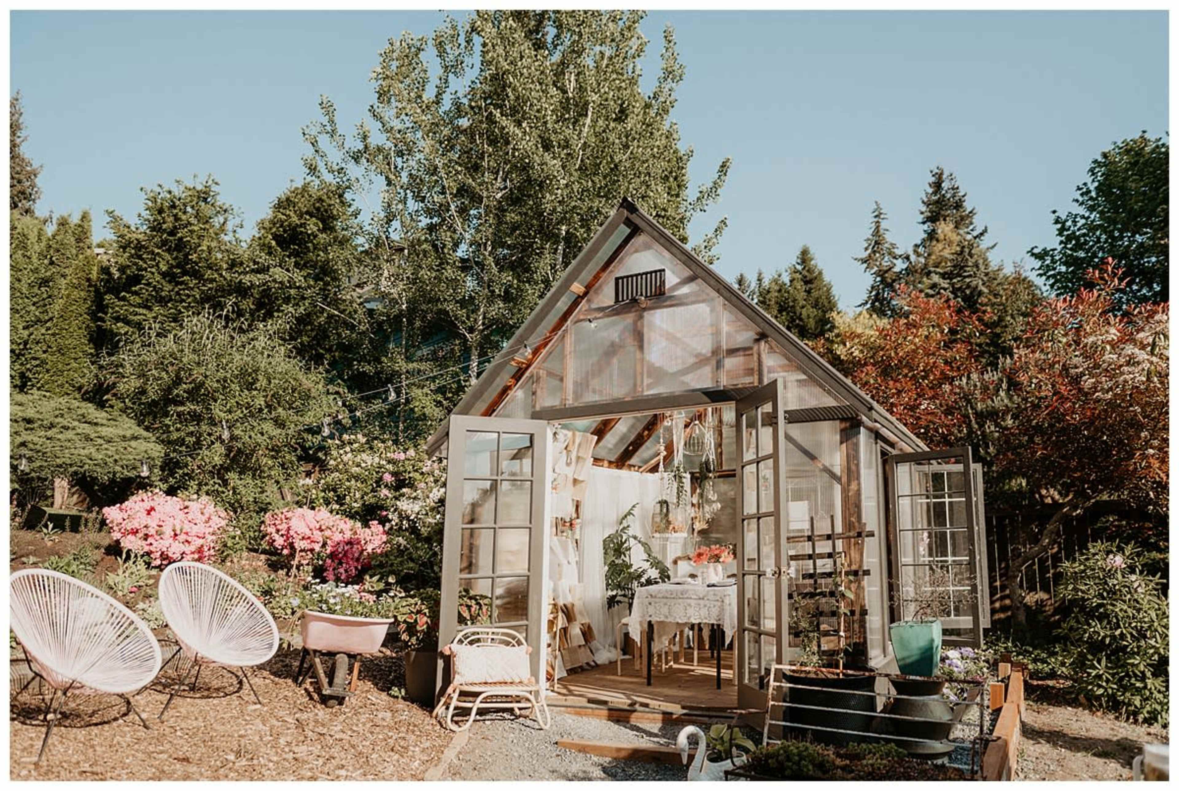 A glass greenhouse with open double doors is surrounded by greenery and flowering plants, featuring outdoor seating in front.