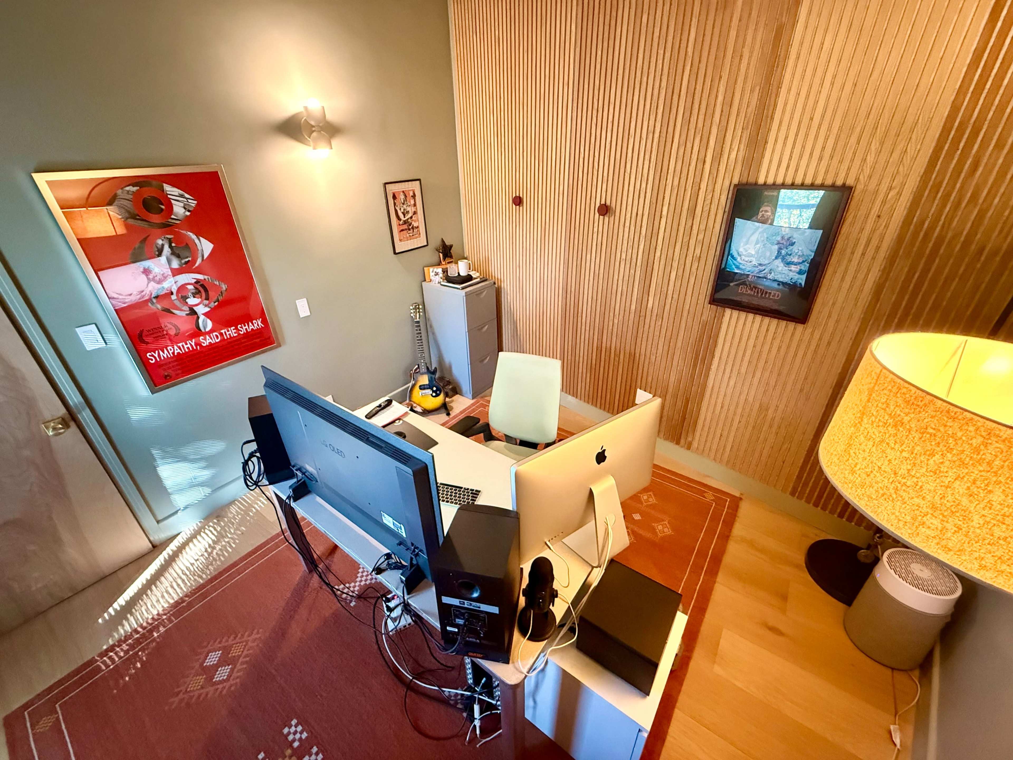 The image shows a modern home office with a desk featuring two computer screens, a guitar, a lamp, and wooden paneling on the walls.