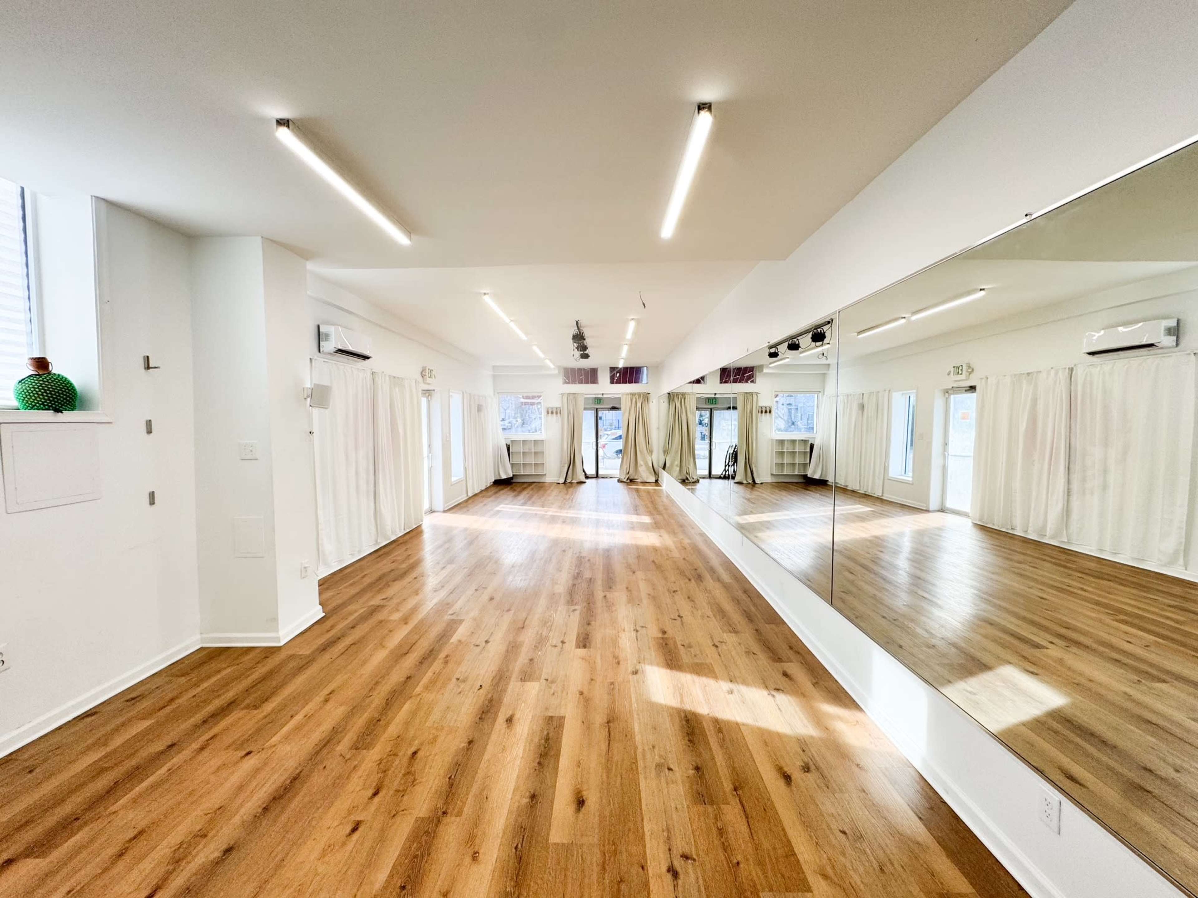 The image shows a spacious, well-lit studio with large mirrors on one wall and a wooden floor, complemented by soft white curtains.