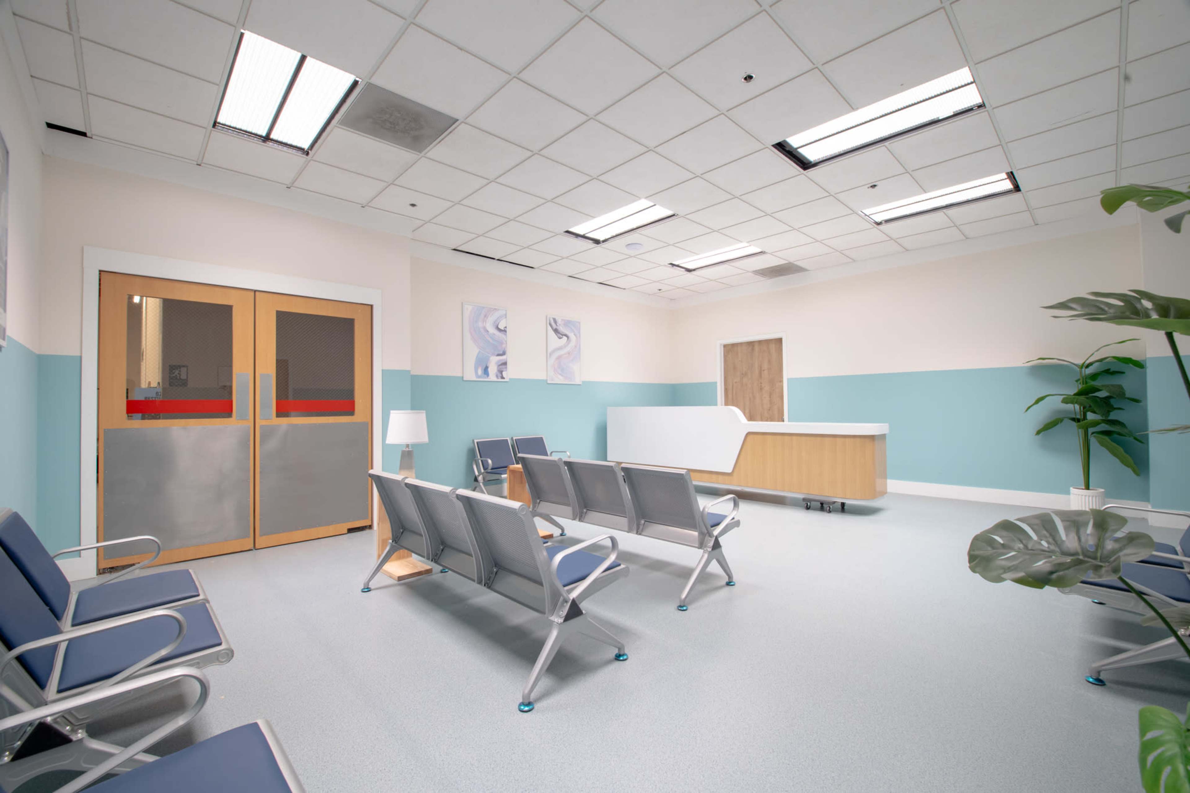 The image shows a clean, well-lit waiting area in a healthcare facility, featuring several gray chairs, a reception desk, and a potted plant.