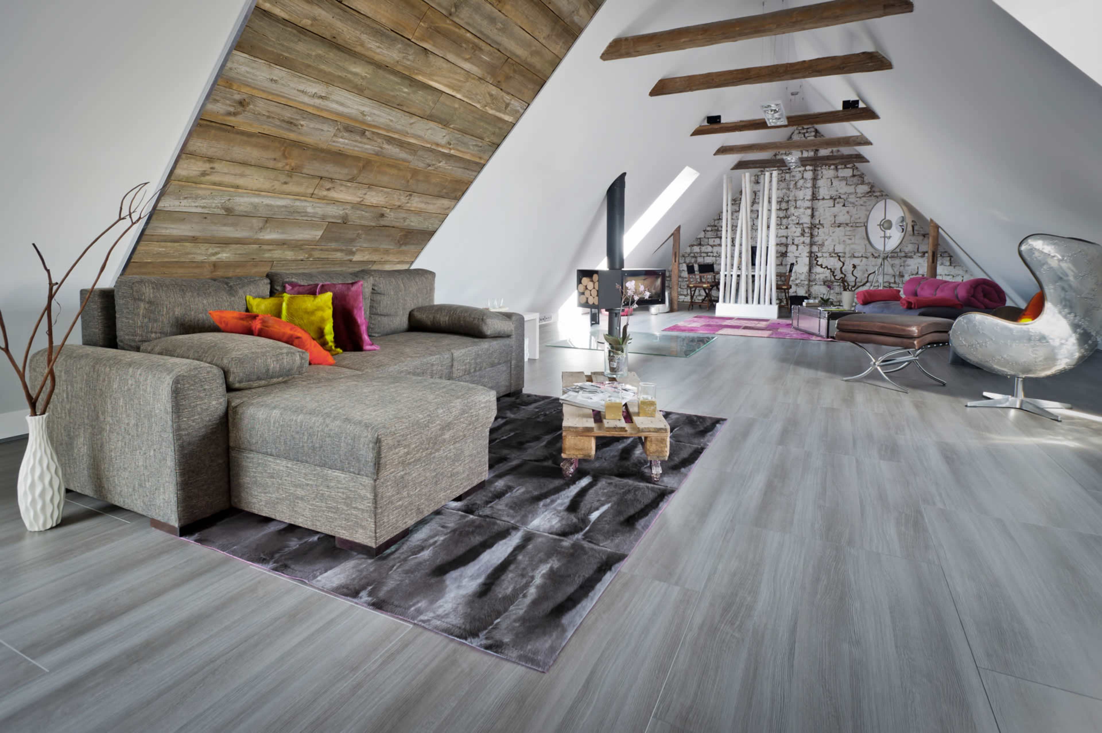 The image shows a modern attic living space featuring a gray sofa, a wooden ceiling with exposed beams, and a mix of furniture including a decorative chair and a television.