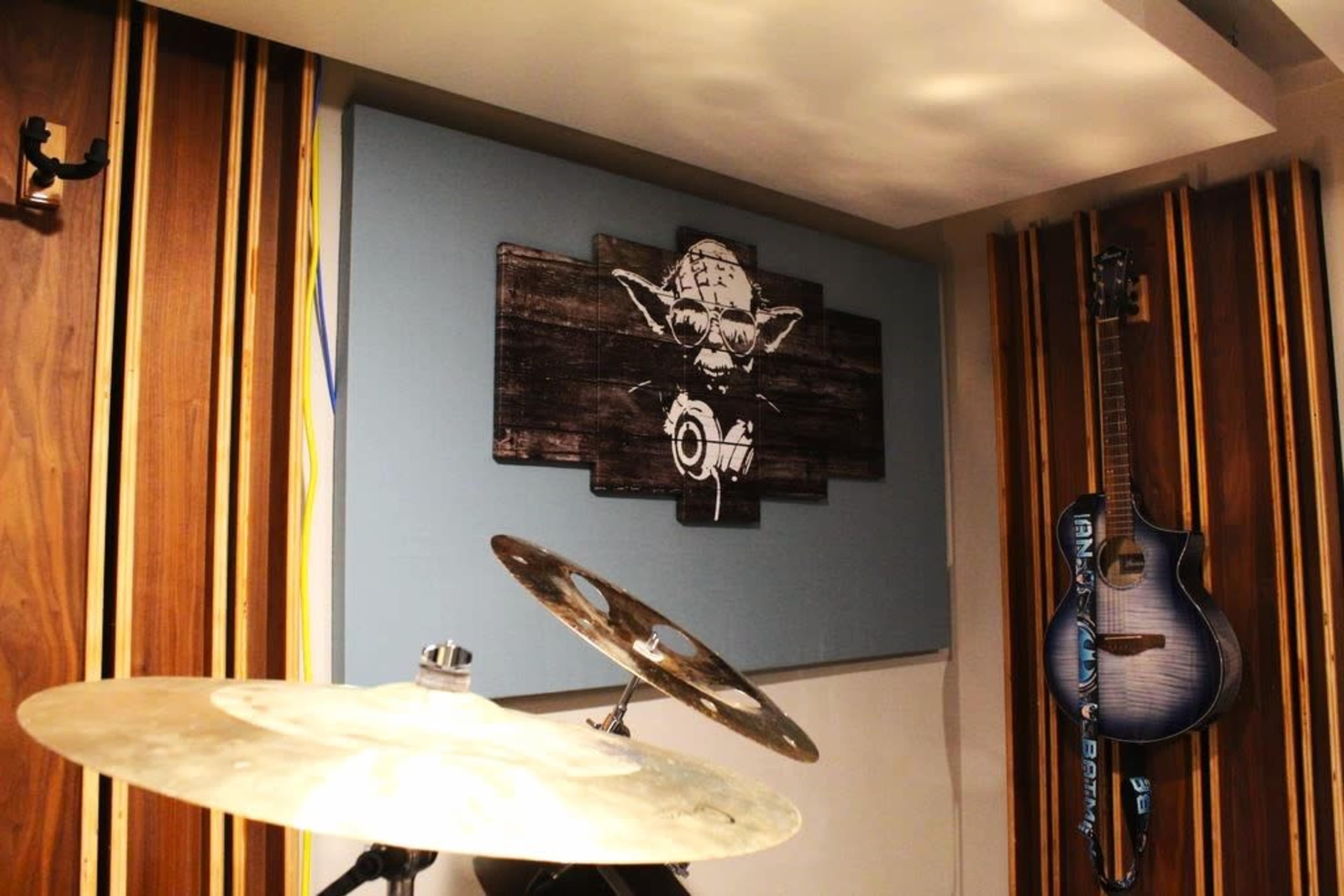 The image shows a music studio with a drum set in the foreground, a graphic of a creature mounted on a wooden panel in the background, and an electric guitar hanging on the wall.