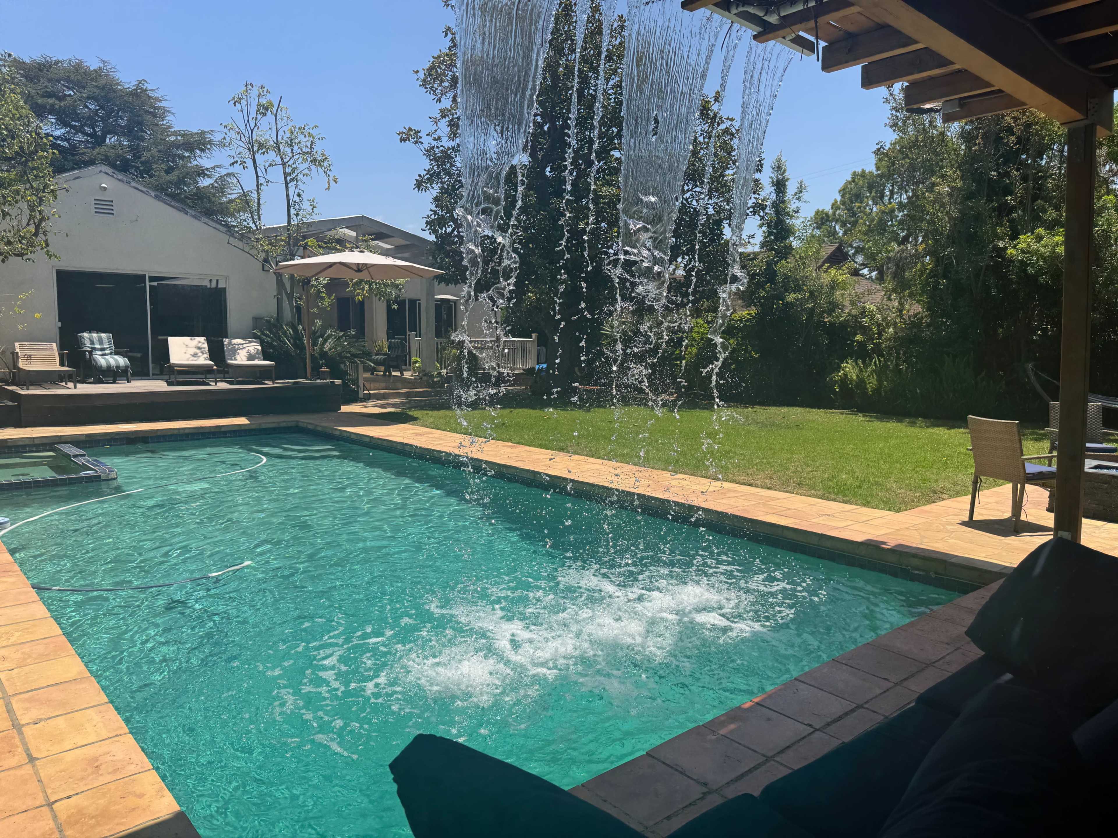 A pool with cascading water features is surrounded by a patio area and lush greenery in a residential backyard.