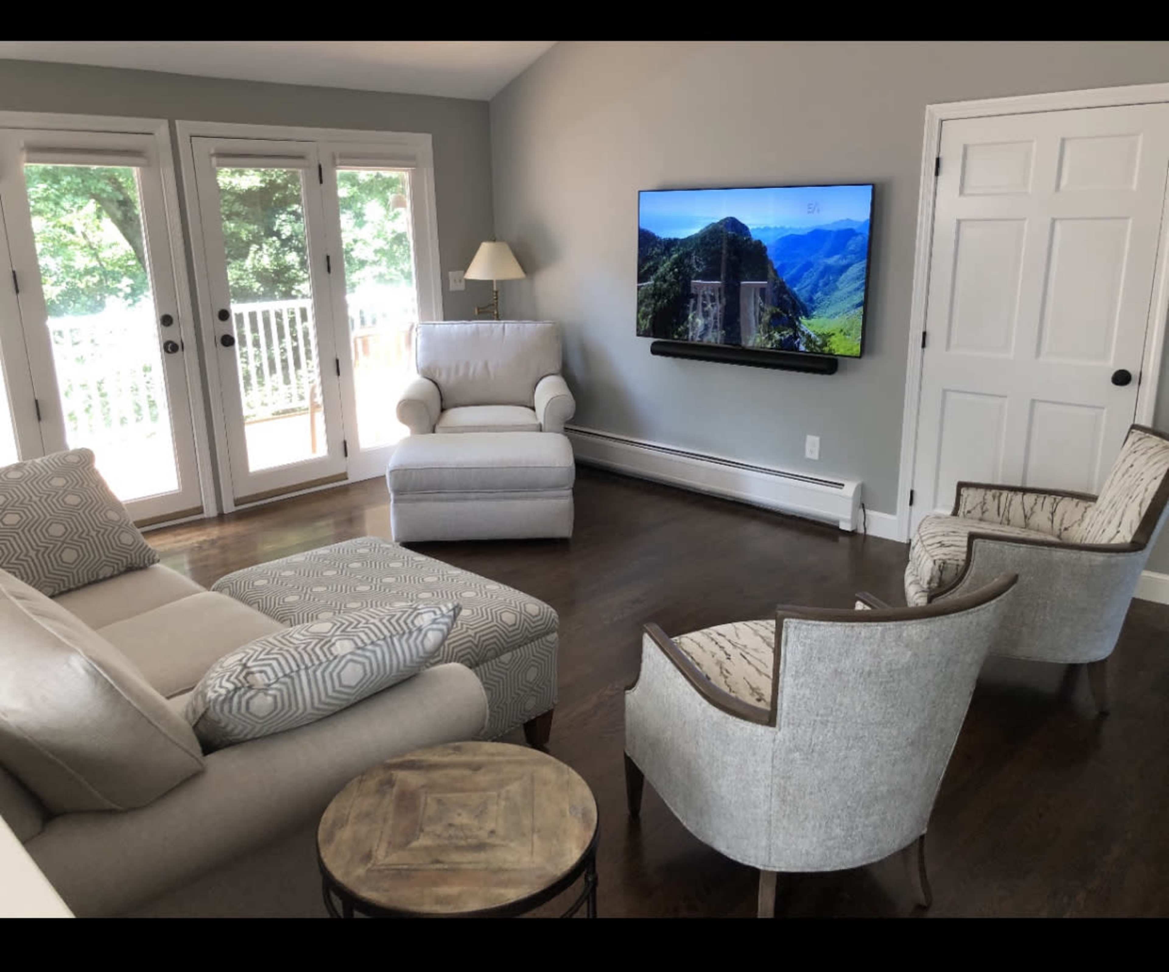 A living room furnished with a light-colored armchair, two upholstered chairs, a sectional sofa, a round table, and a wall-mounted television, with large windows allowing natural light.