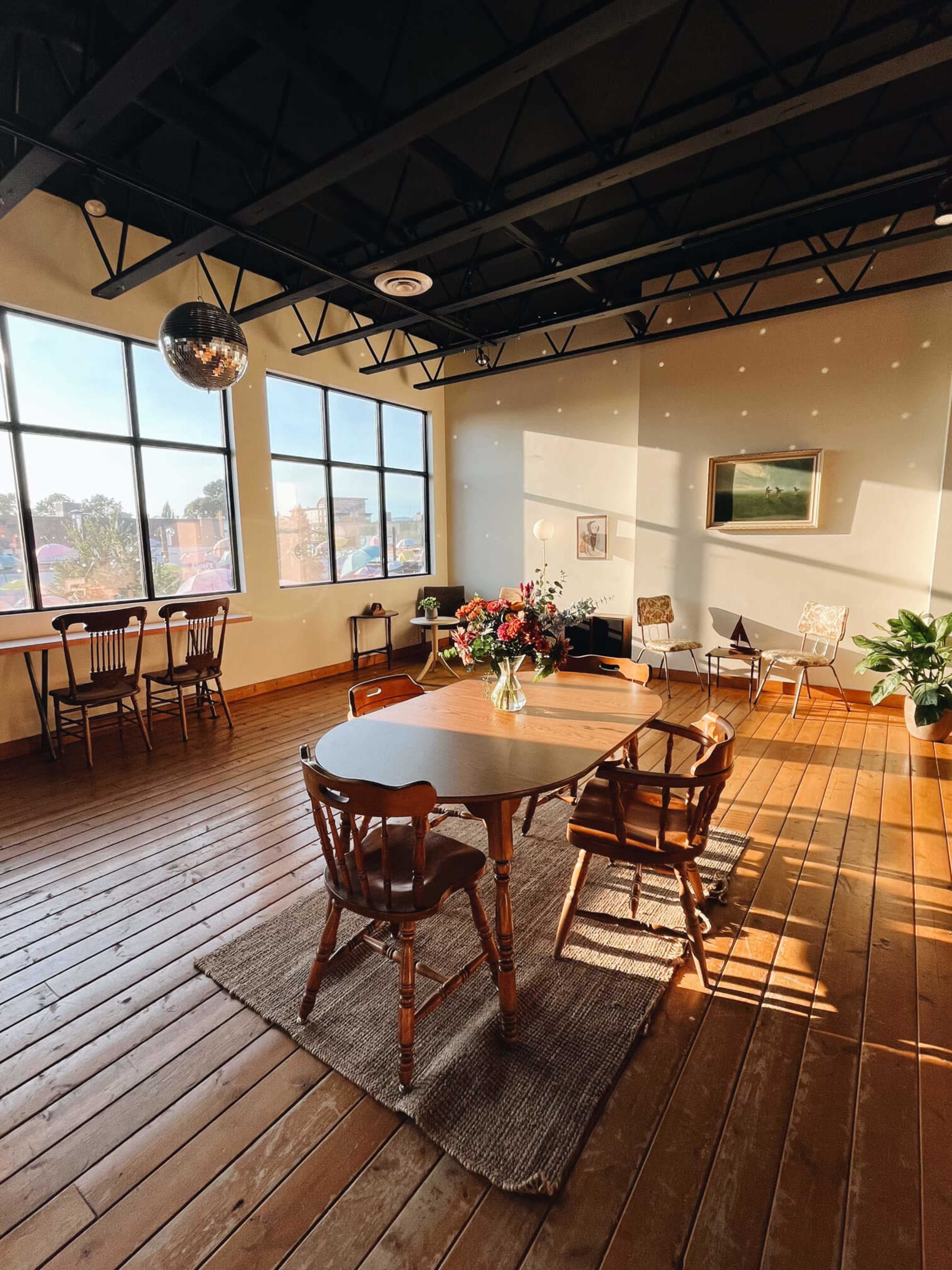 The image shows a spacious room with wooden floors, large windows letting in natural light, and a round table surrounded by chairs, adorned with a floral arrangement.