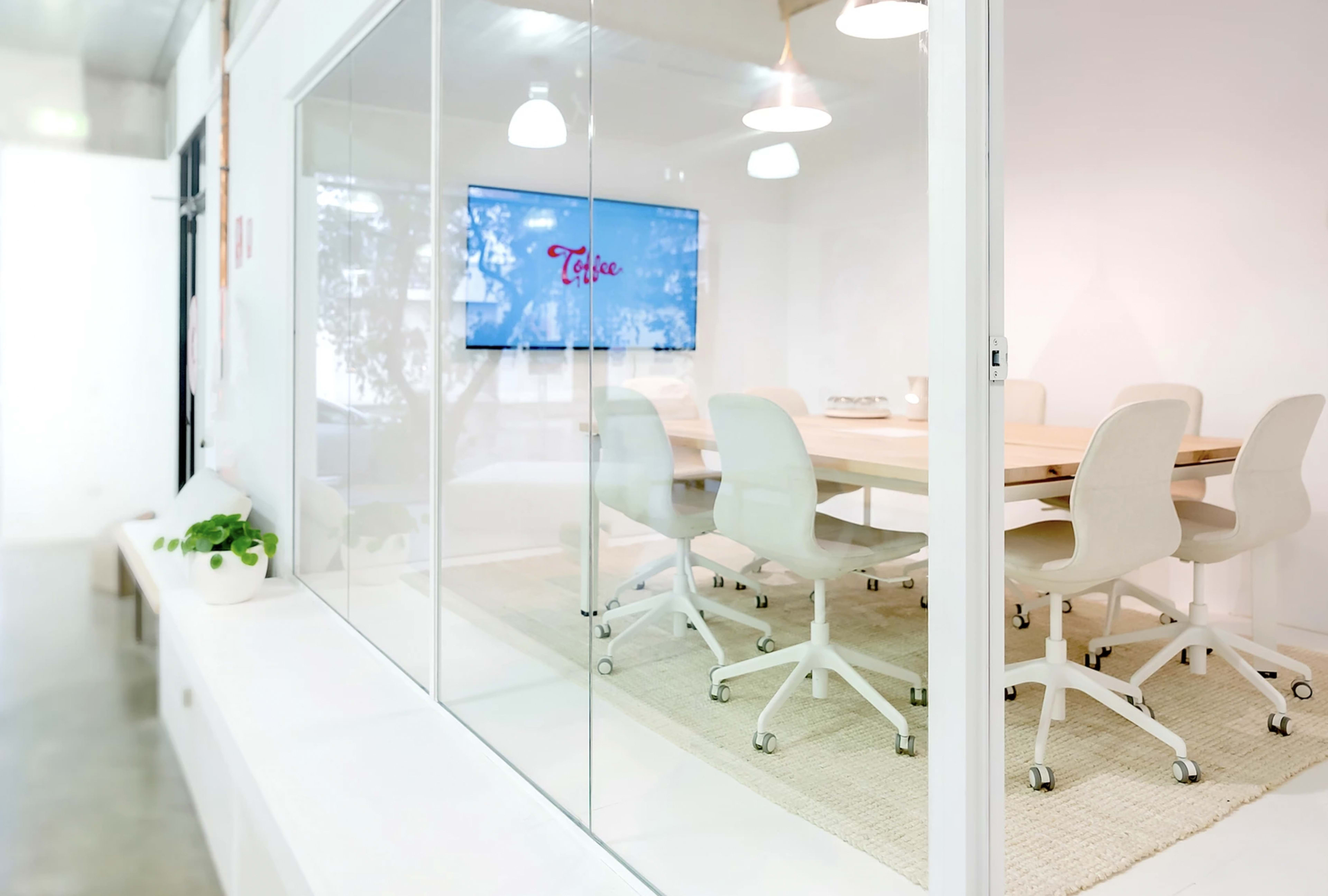 A glass-walled conference room features a large wooden table surrounded by white chairs, with a television displaying the word "Talis" in the background.