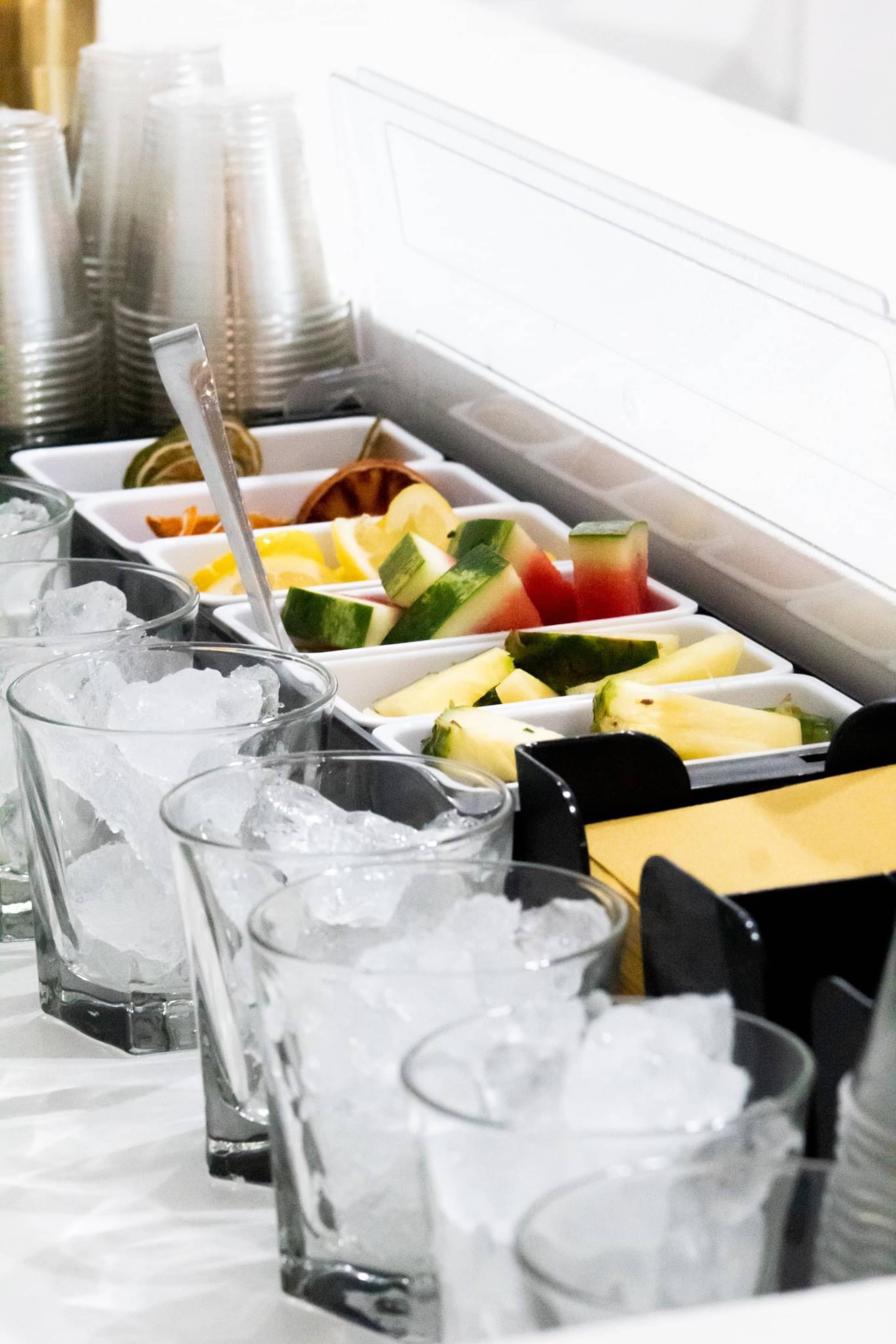 A display of ice-filled glasses is arranged beside bowls of sliced fruits, including watermelon, pineapple, and citrus, in a serving area.
