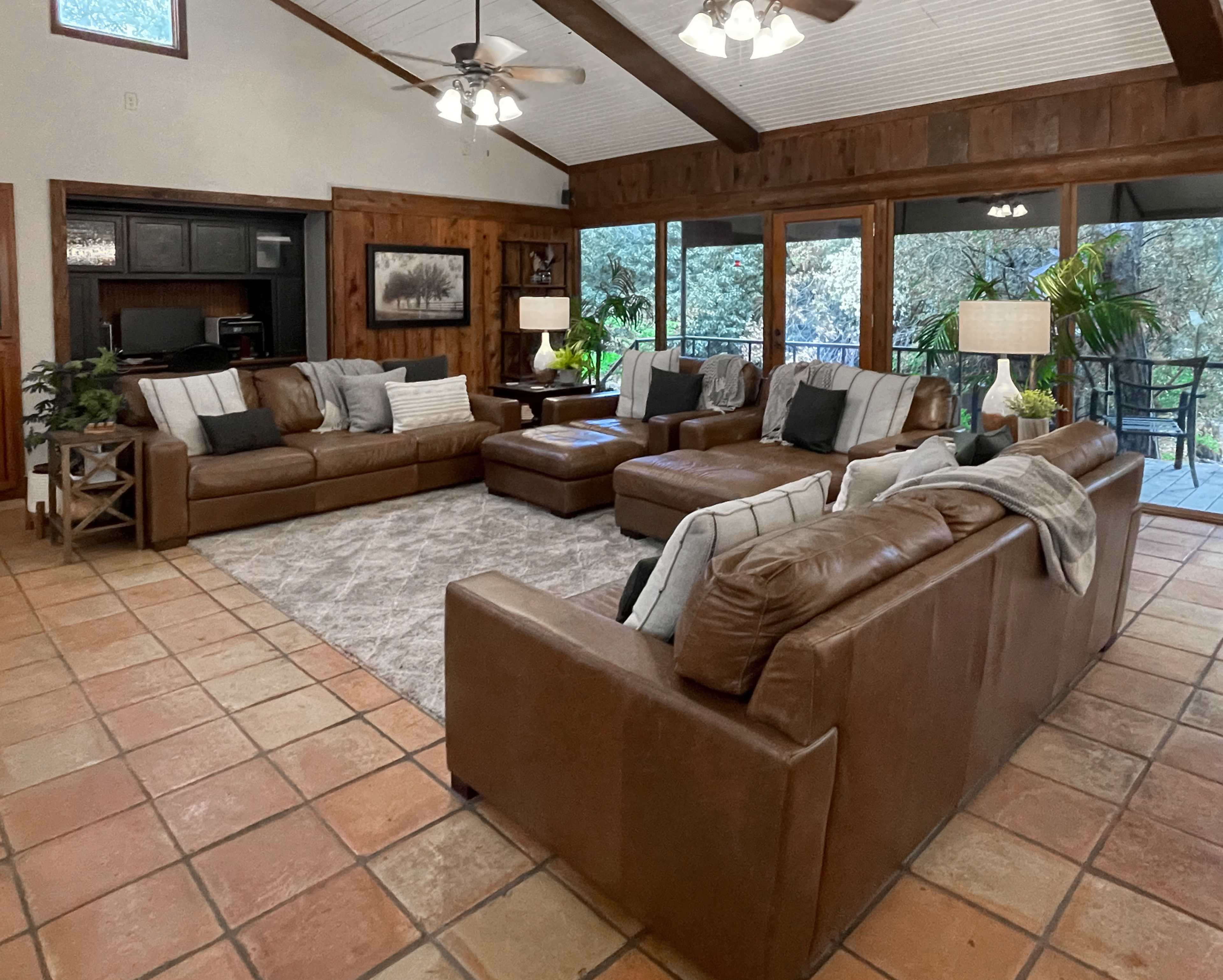 The spacious living room features brown leather sofas arranged around a central area rug, illuminated by ceiling fans and large windows displaying a view of the outdoors.