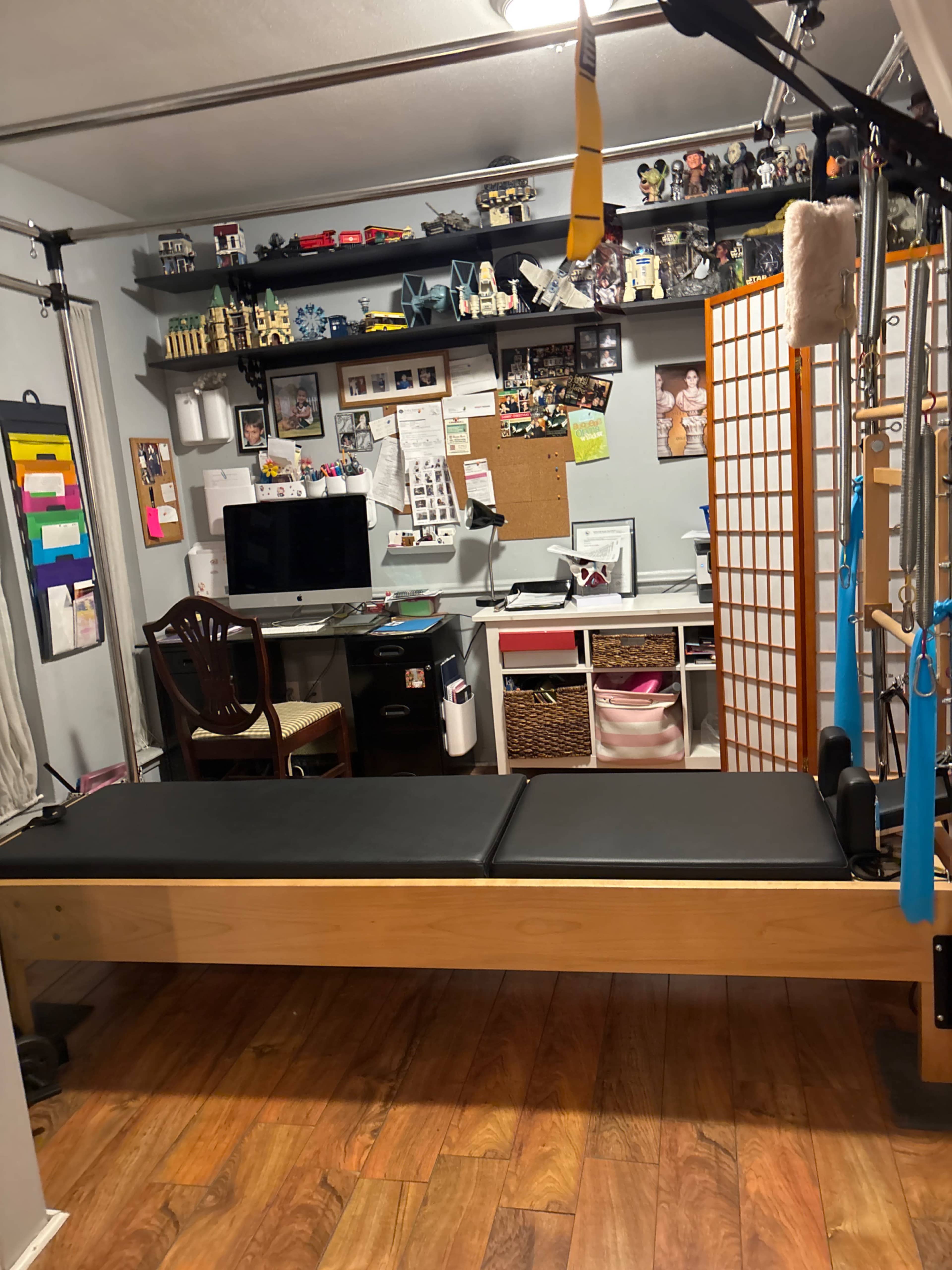 The image shows a Pilates reformer in a home studio with a desk and various items on the wall, including a bulletin board and toys on shelves.