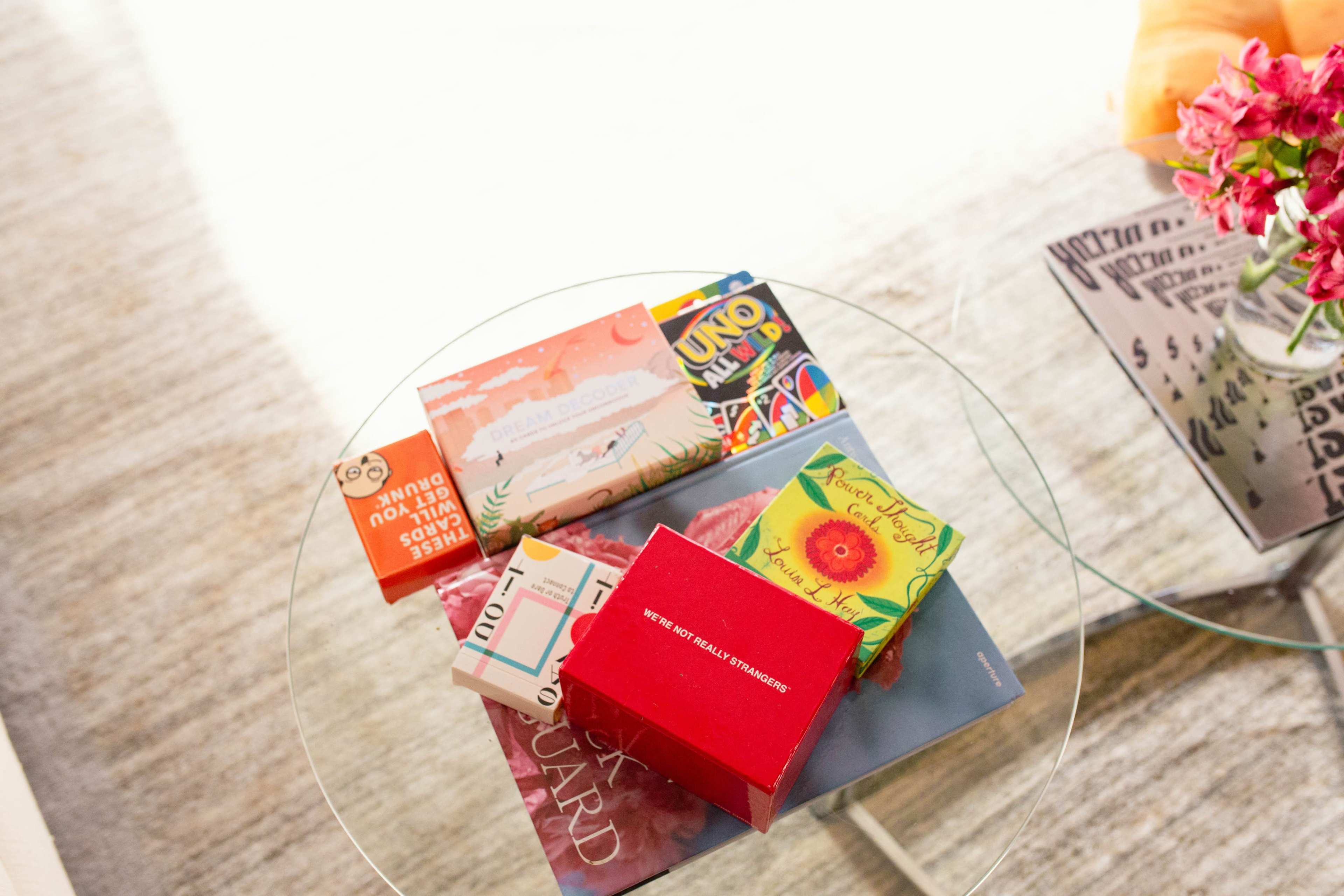 A glass table displays various books and a box, with a bouquet of flowers in the background.