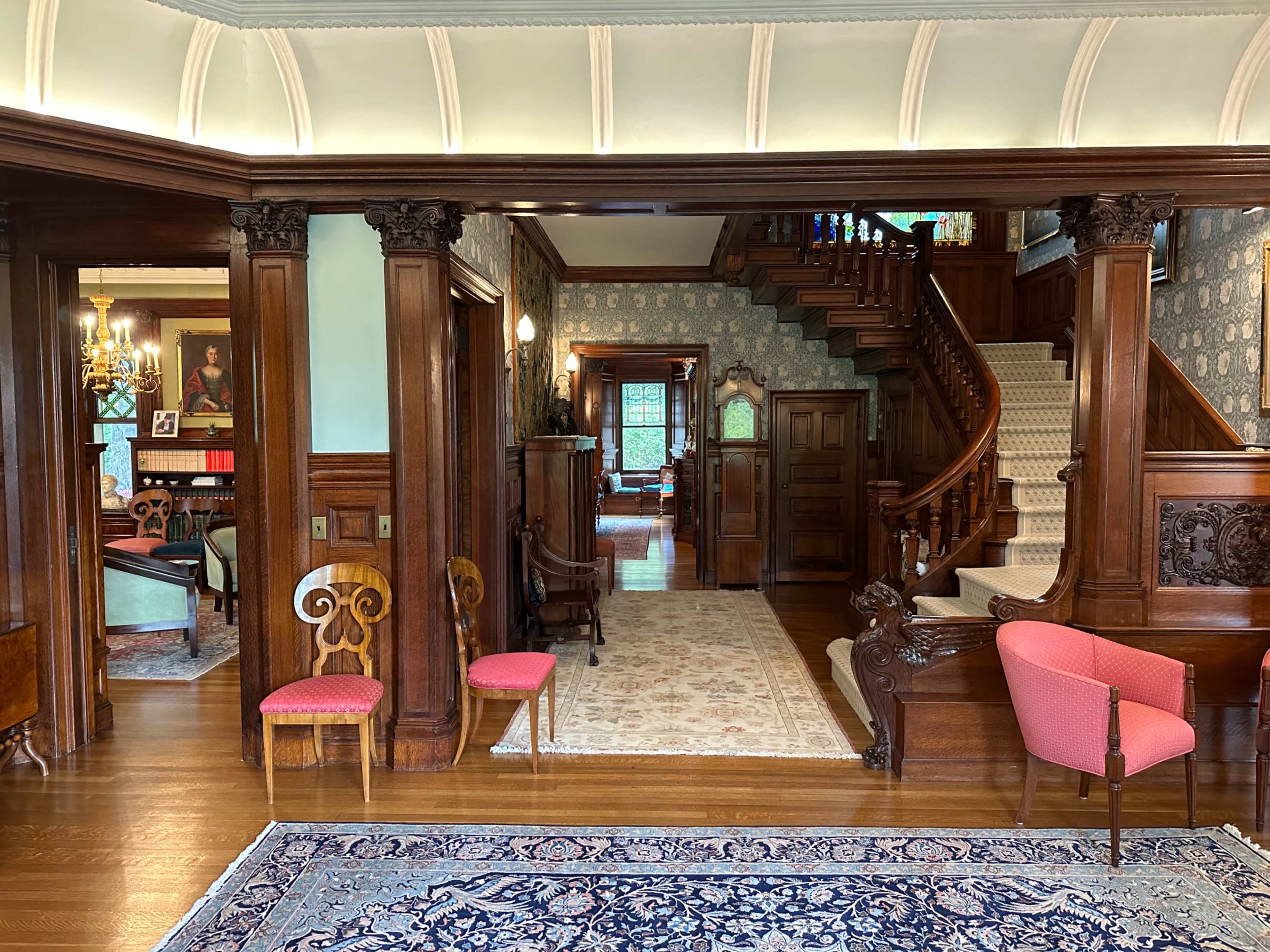 The image shows an elegantly designed foyer of a historical home, featuring wooden columns, a staircase, and vintage furnishings.