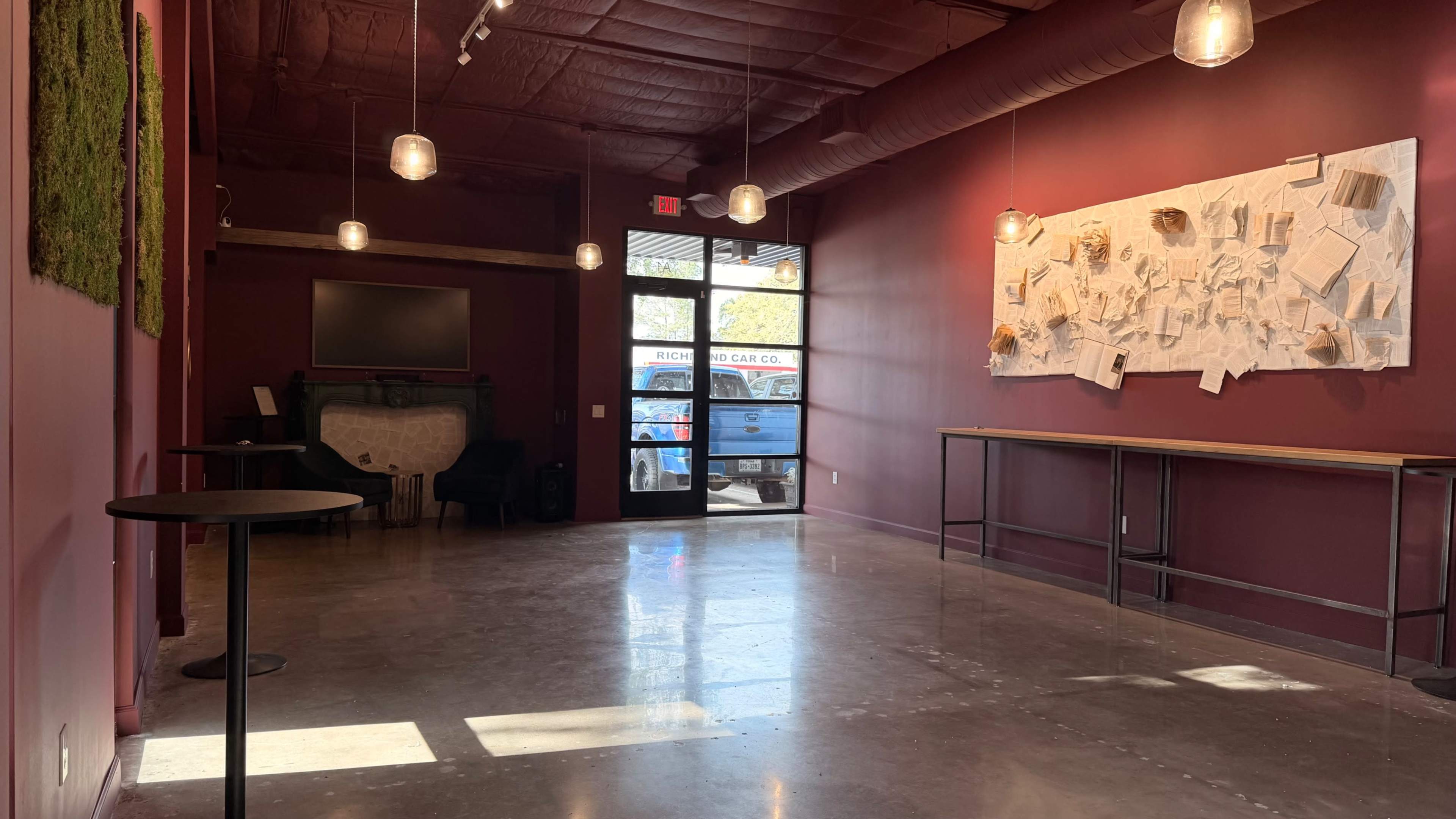 The image shows a minimalistic interior space with a red wall, pendant lights, and a large textured artwork on one wall, leading to an entrance with large glass doors.