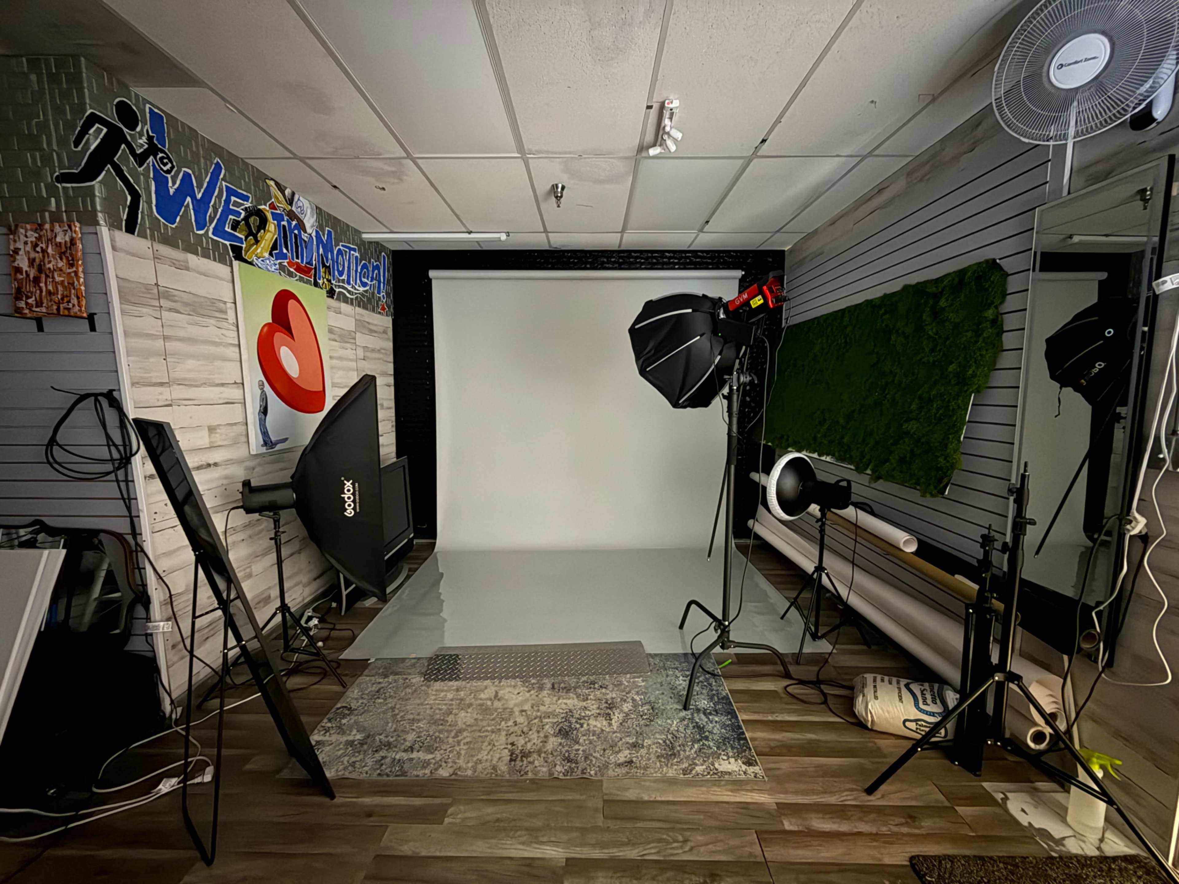 The image shows a photography studio with a plain backdrop, lighting equipment, and various props arranged around the space.