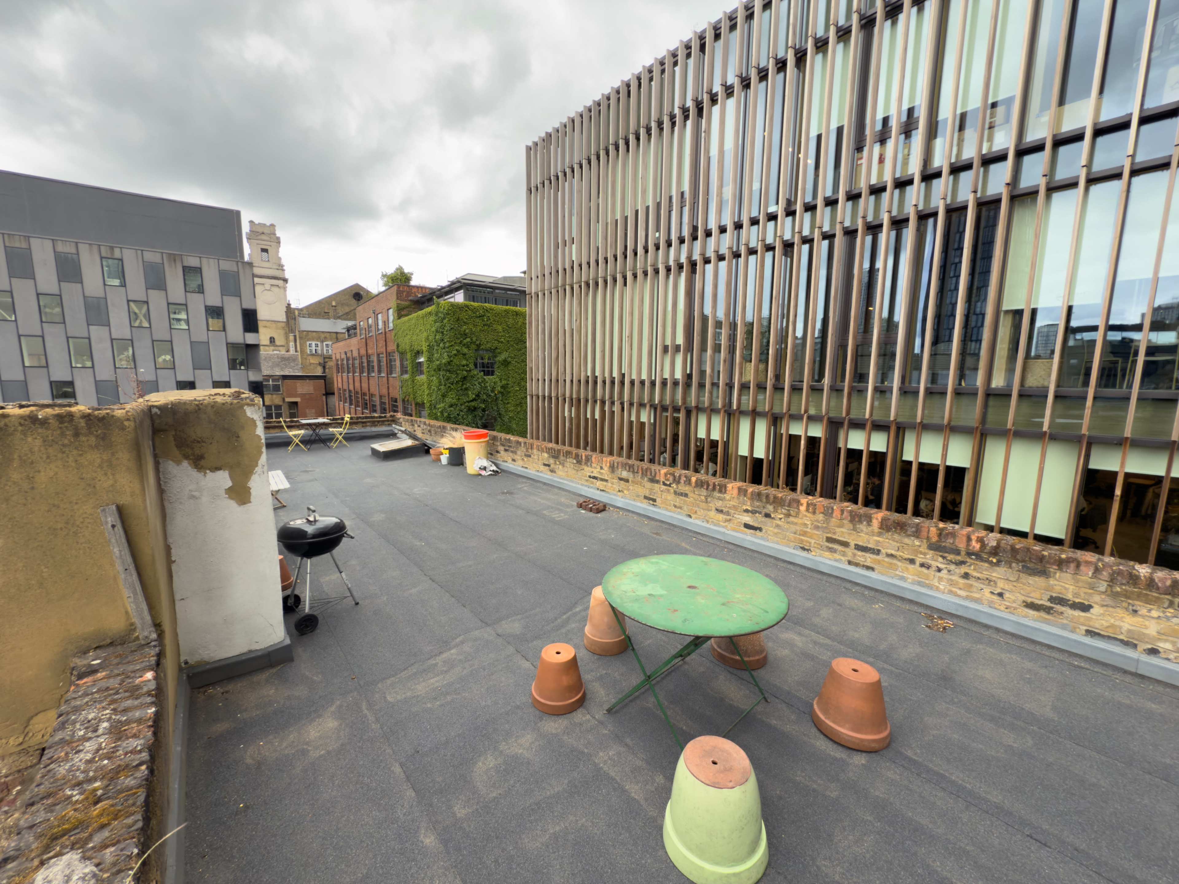 The scene shows a rooftop area with a green table and several terracotta pots scattered on a gray surface, surrounded by modern buildings in the background.