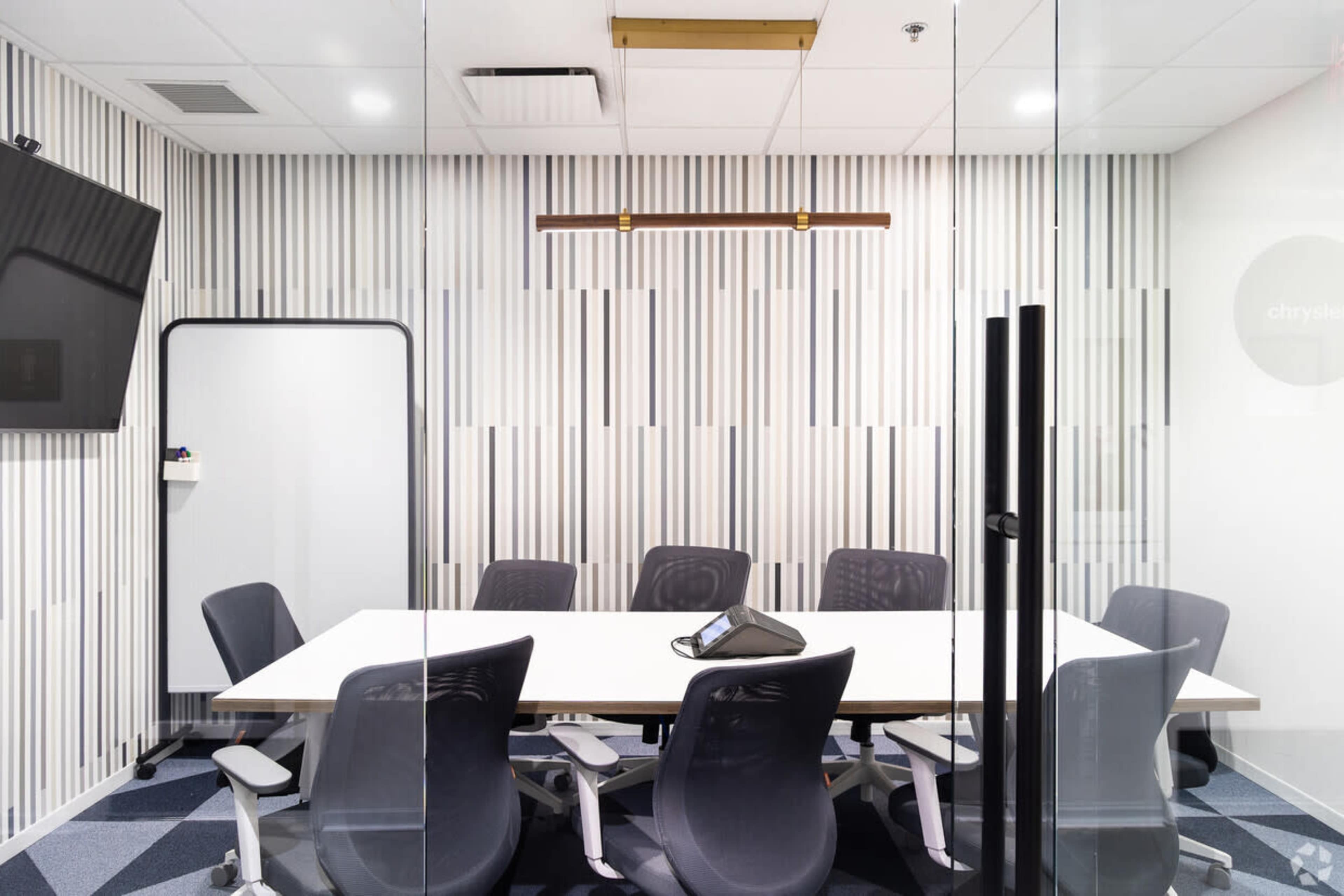 A conference room features a large table surrounded by ergonomic chairs, with a wall-mounted screen and a whiteboard.