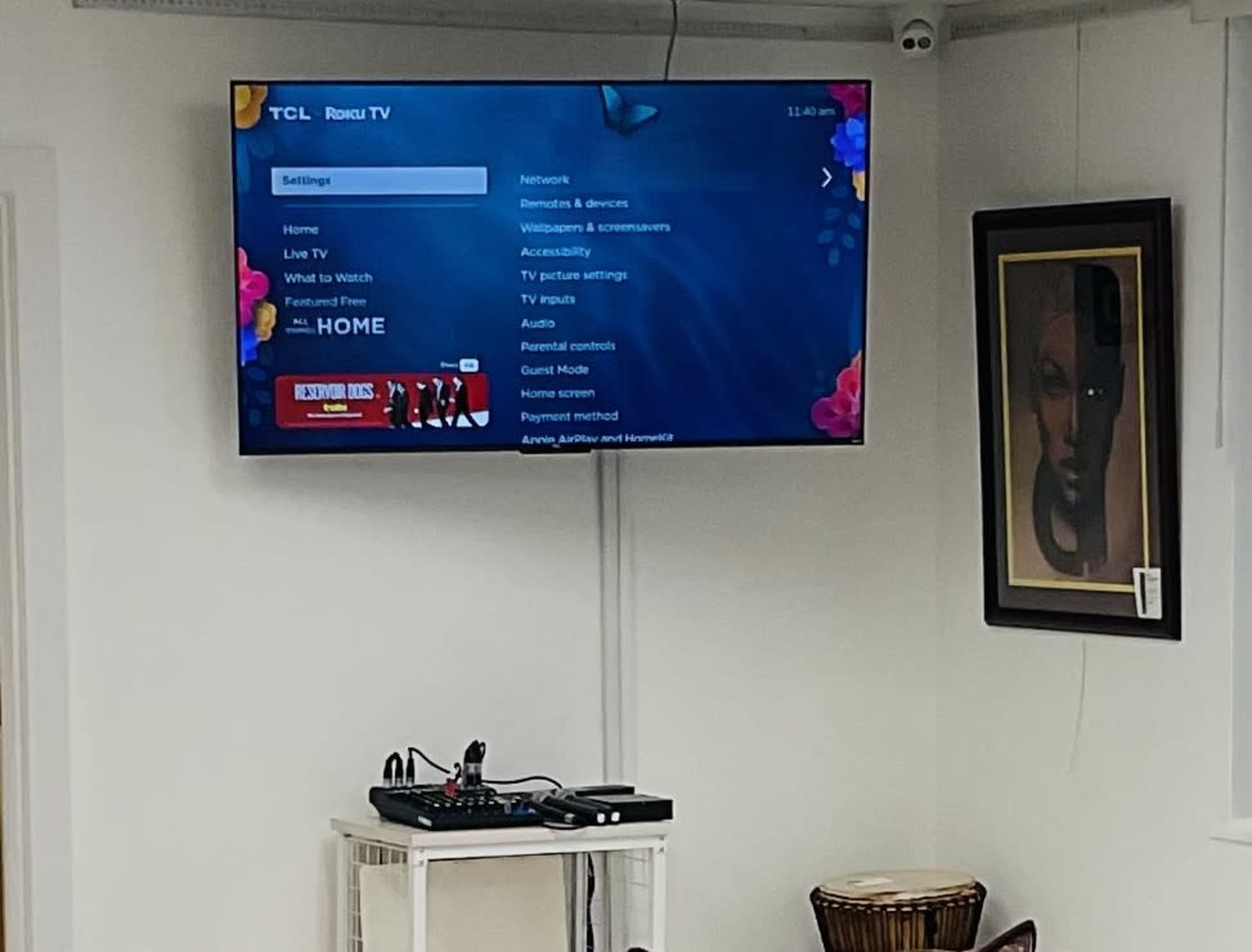A TCL Roku TV displaying the settings menu is mounted on a white wall above a small table with electronic devices and a framed artwork.