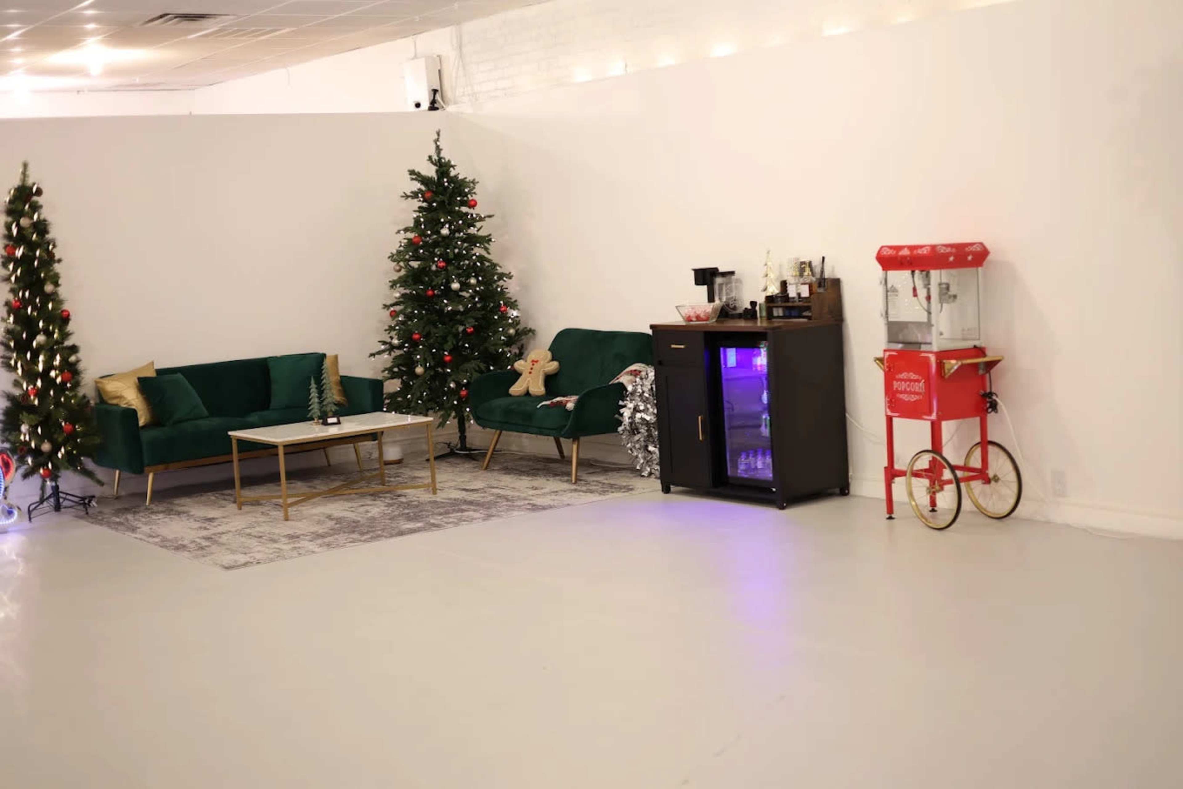 The scene features two decorated Christmas trees, a pair of green sofas with a coffee table, and a vintage popcorn machine against a white wall in a spacious room.