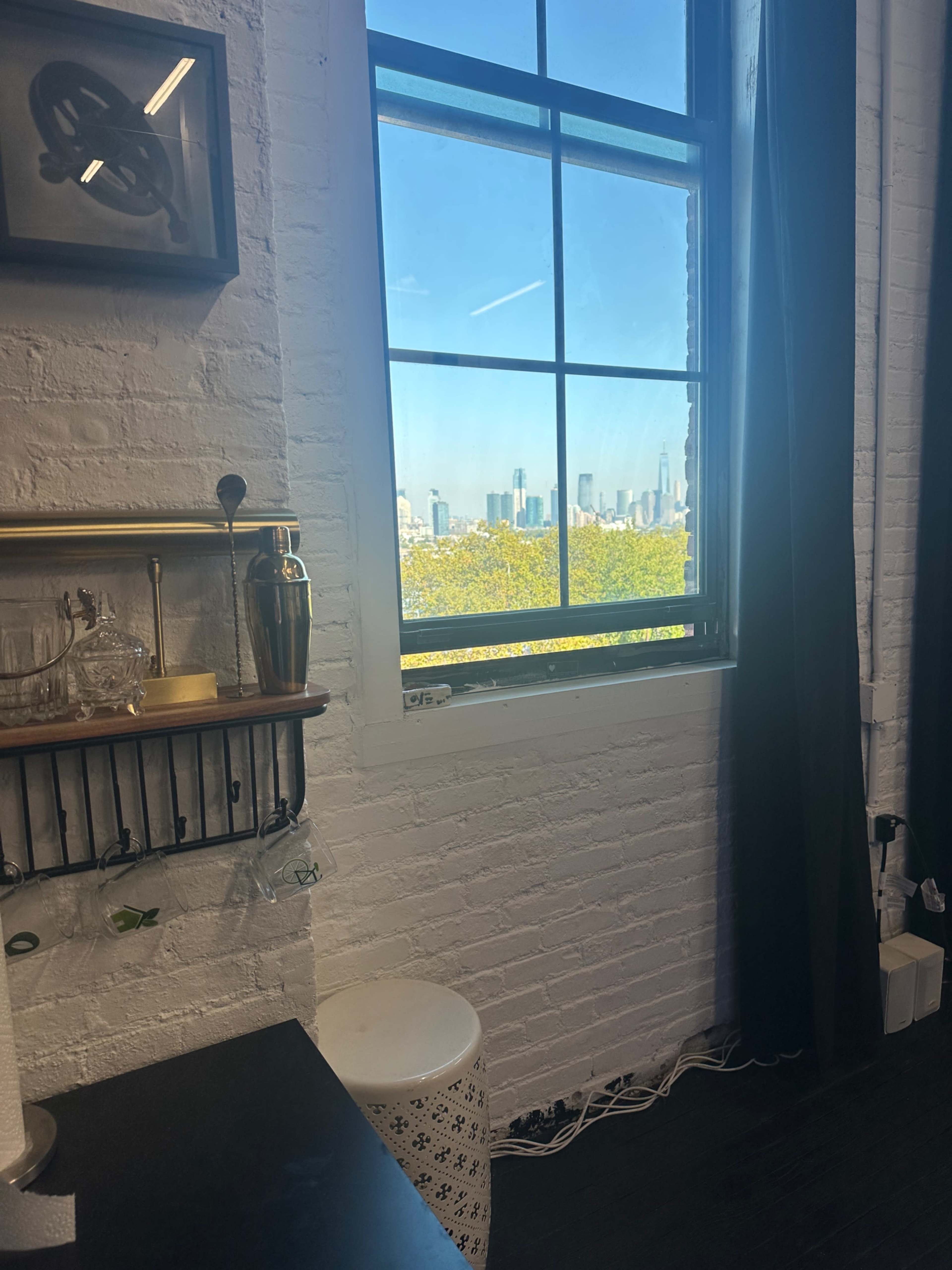 A window with a view of a city skyline is framed by a white-brick wall and dark curtains, with a bar cart visible nearby.