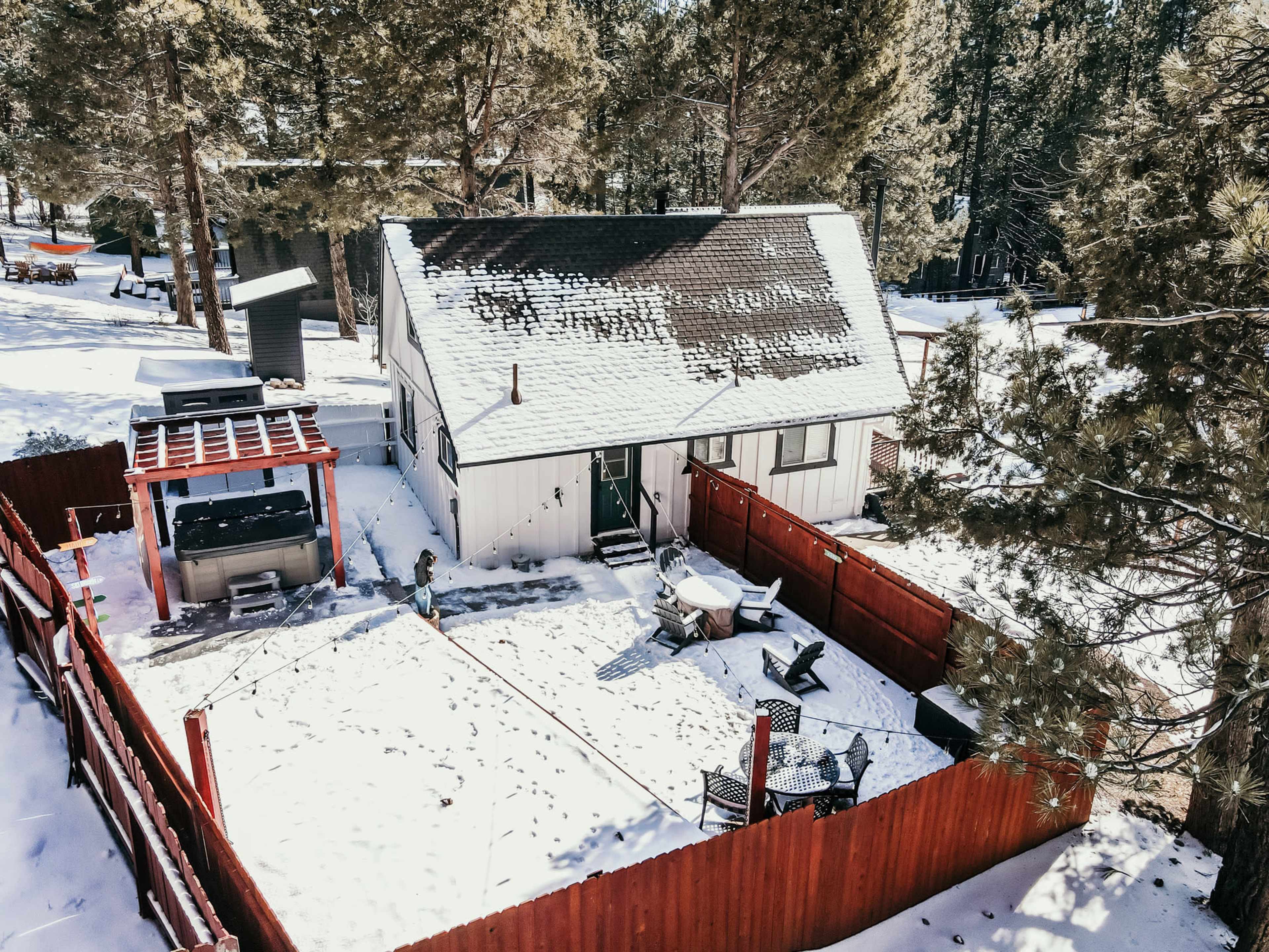 A small, snow-covered house with a fenced yard and outdoor seating area is surrounded by tall pine trees.