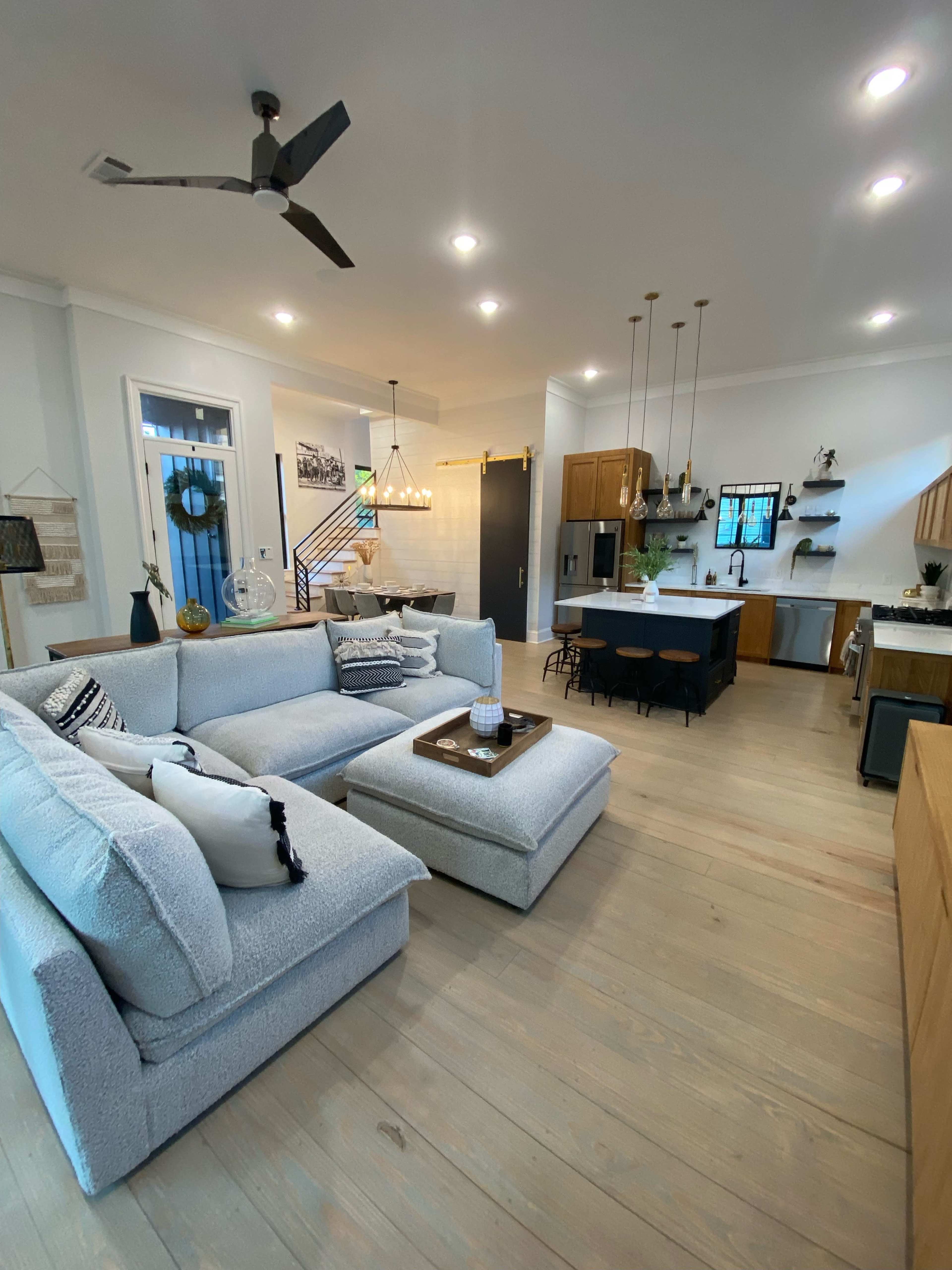 A modern living room with a light-colored sectional sofa, a coffee table, and an open kitchen area featuring wooden cabinets and a dining table.