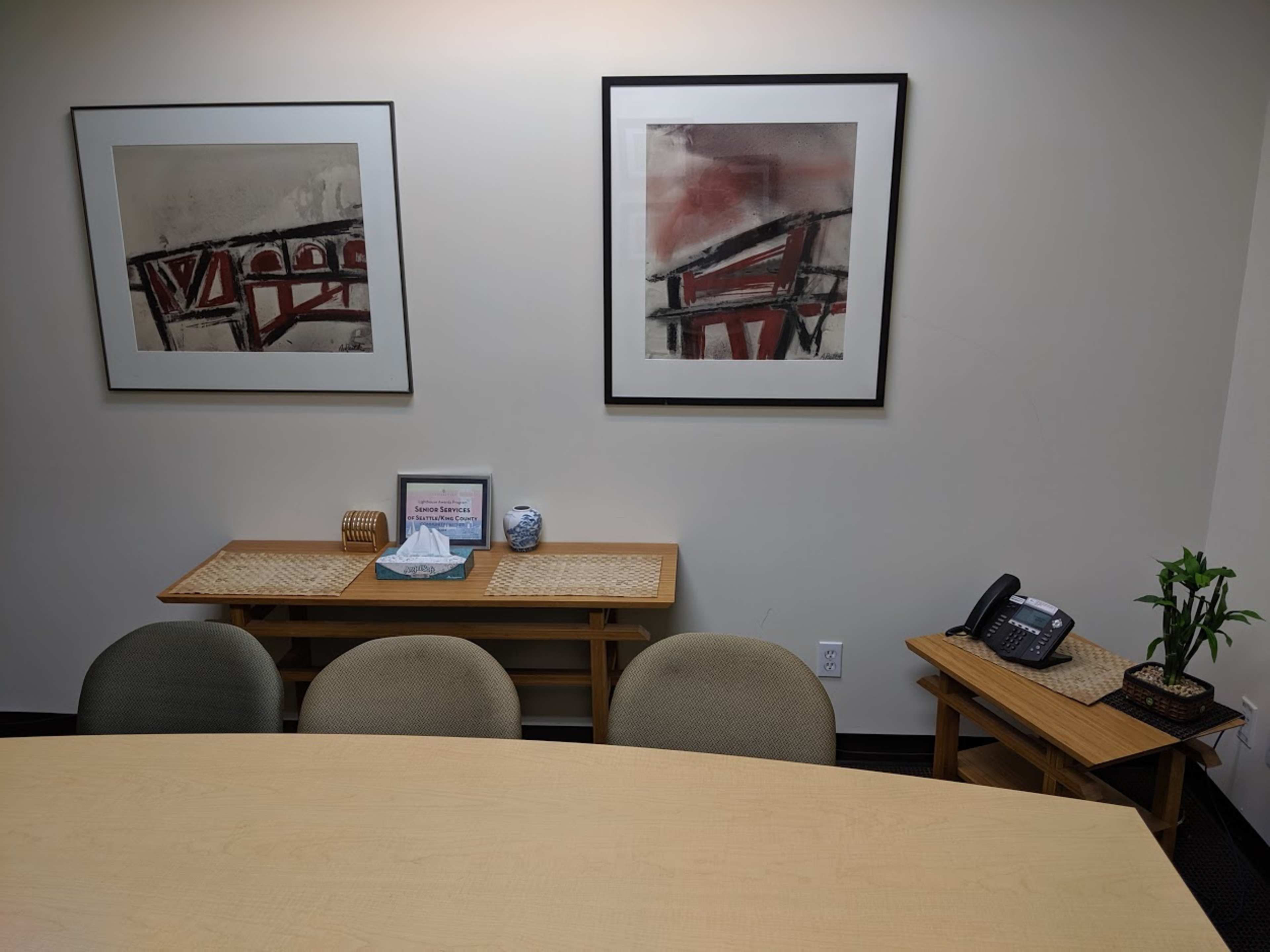 The image shows a small conference room with a wooden table, three chairs, a phone on a side table, and two abstract wall art pieces.