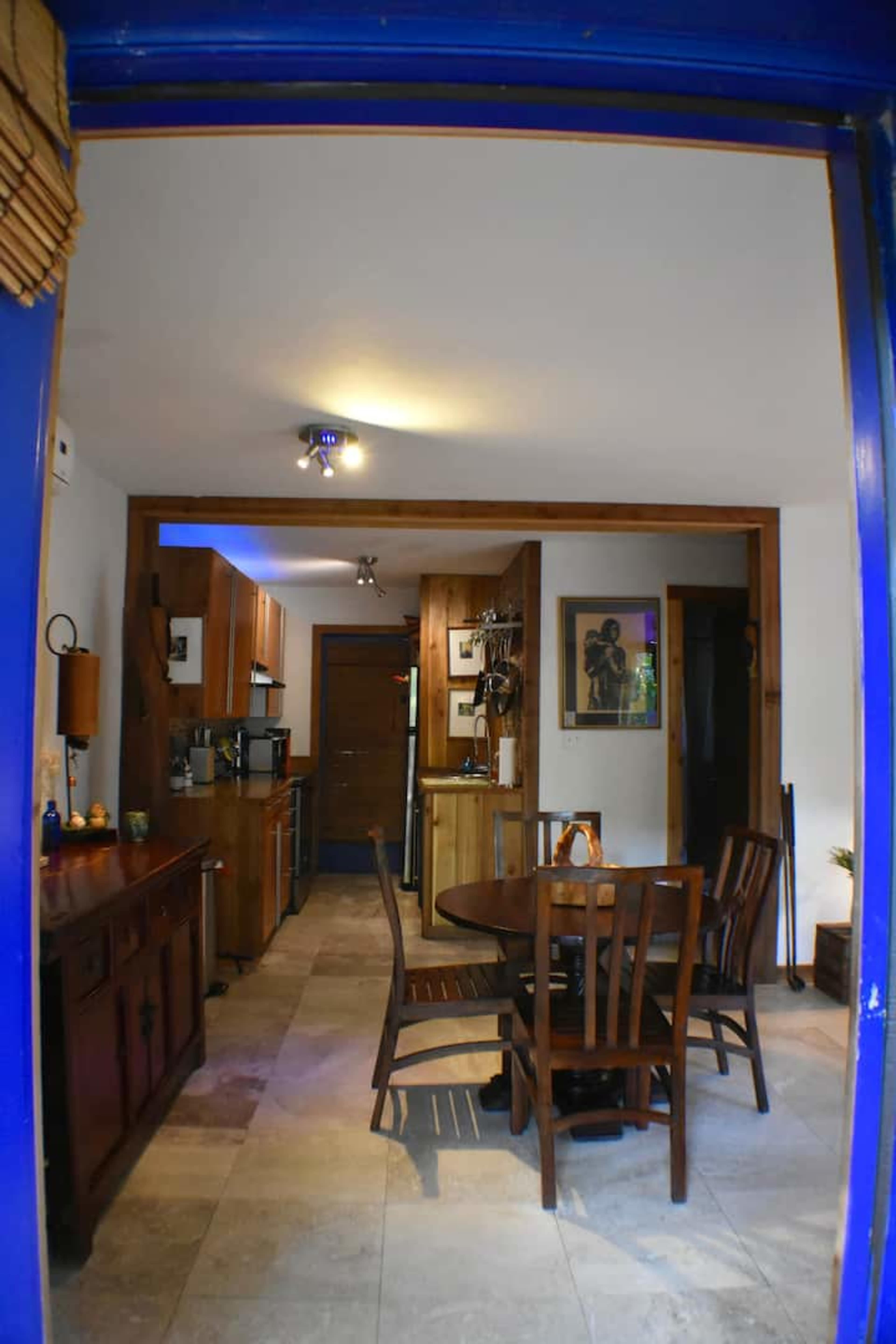 The image shows a kitchen with wooden cabinets, a circular dining table, and tiled flooring, viewed through a blue-framed doorway.