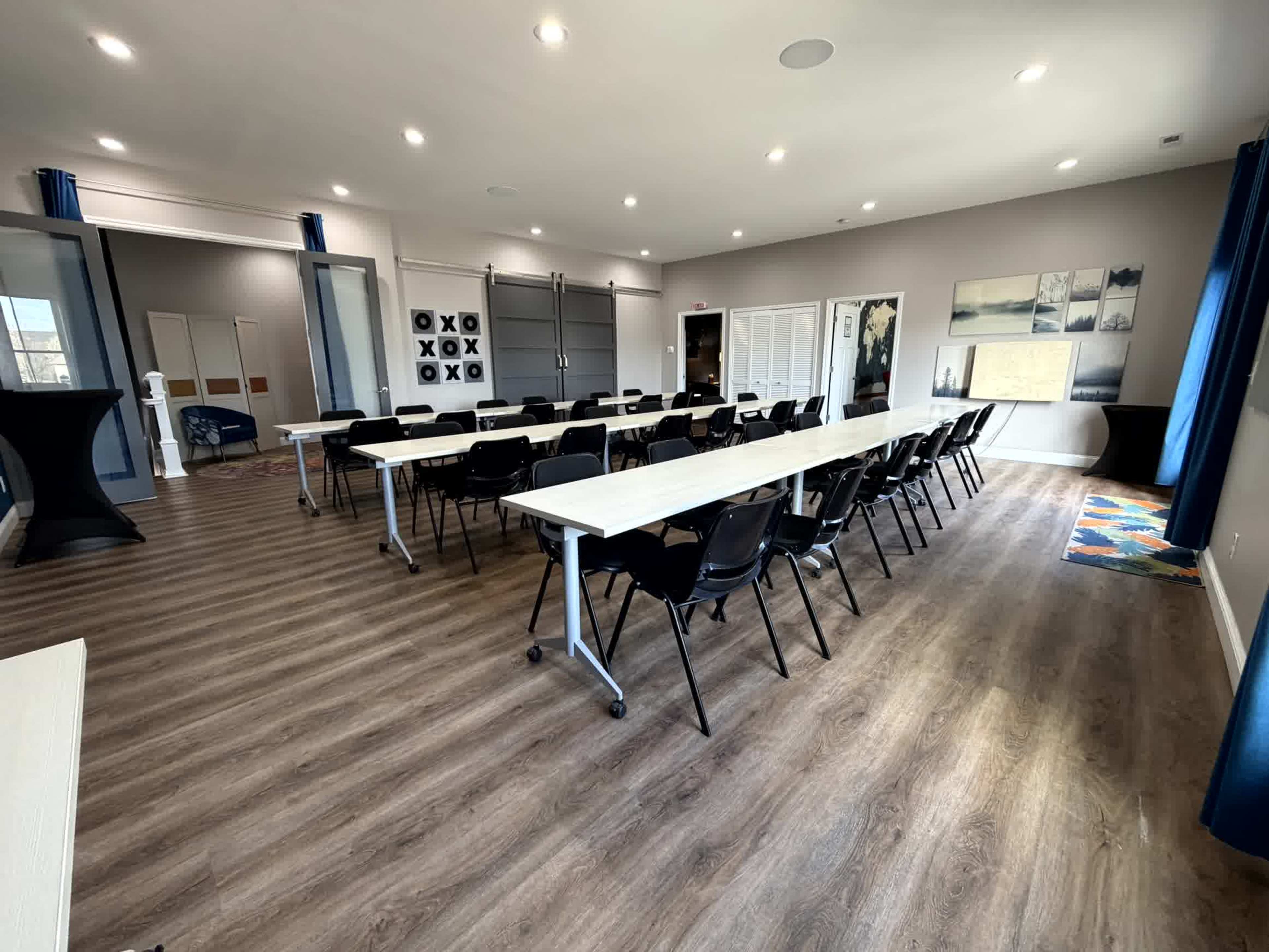 The image shows a conference room set up with rows of tables and black chairs, featuring modern decor and bright lighting.
